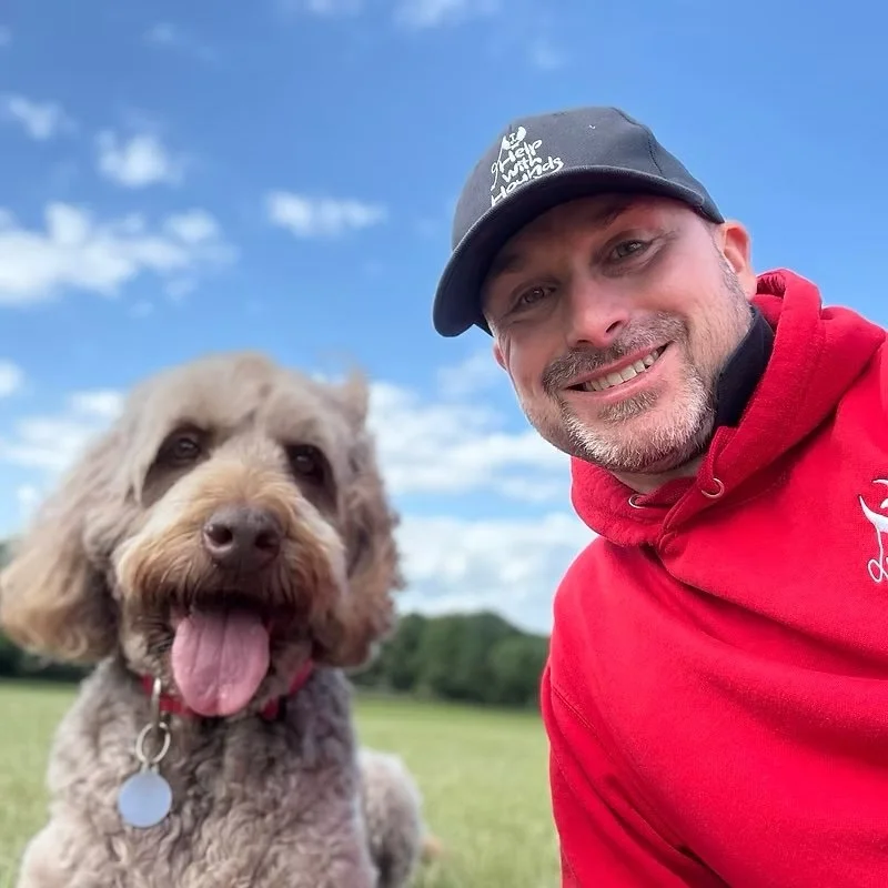 A dog trainer with his dog taking a selfie outdoors on a sunny day with a blue sky and some clouds in the background. The man is smiling, wearing a red hoodie and a black cap, and the dog has its tongue out, looking happy.