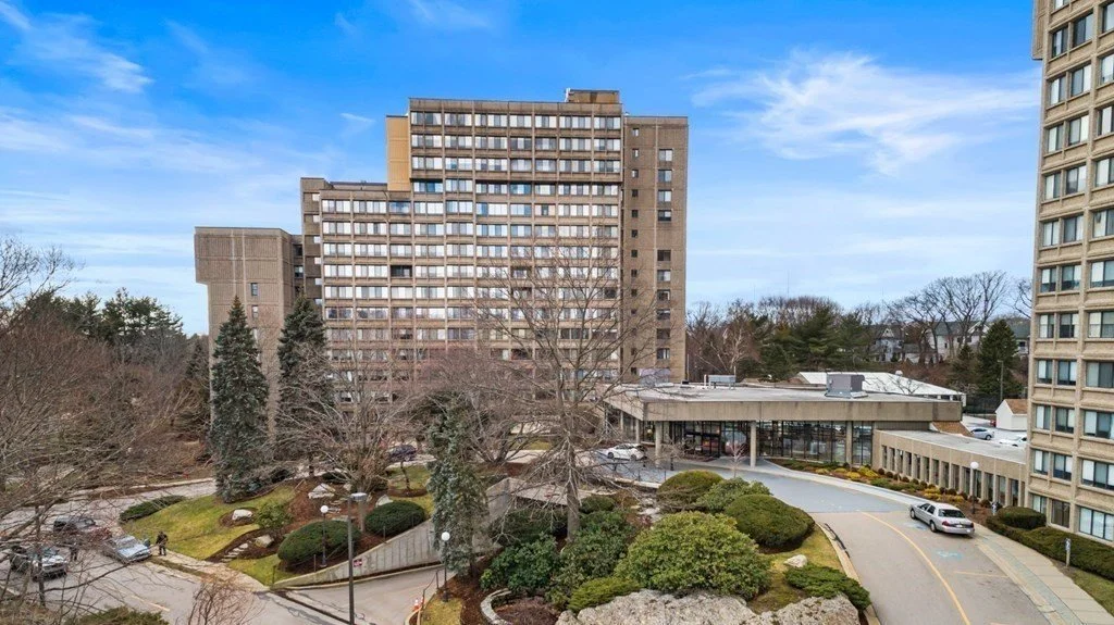 A view of a residential building complex with high-rise apartments, trees, and a parking area, under a partly cloudy sky.