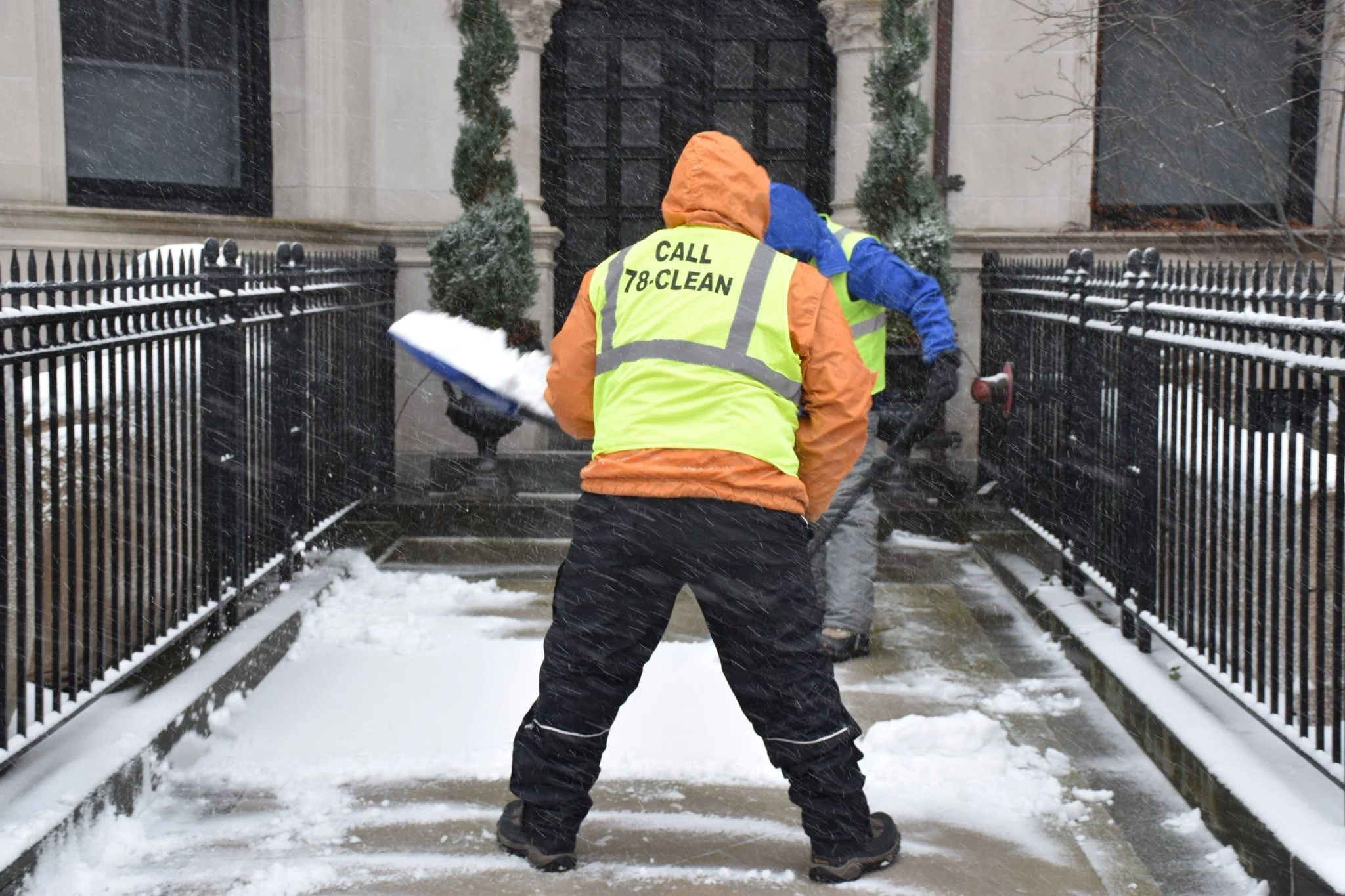 A worker in a yellow safety vest with the text 'CALL 78-CLEAN' on the back shoveling snow in front of a house.
