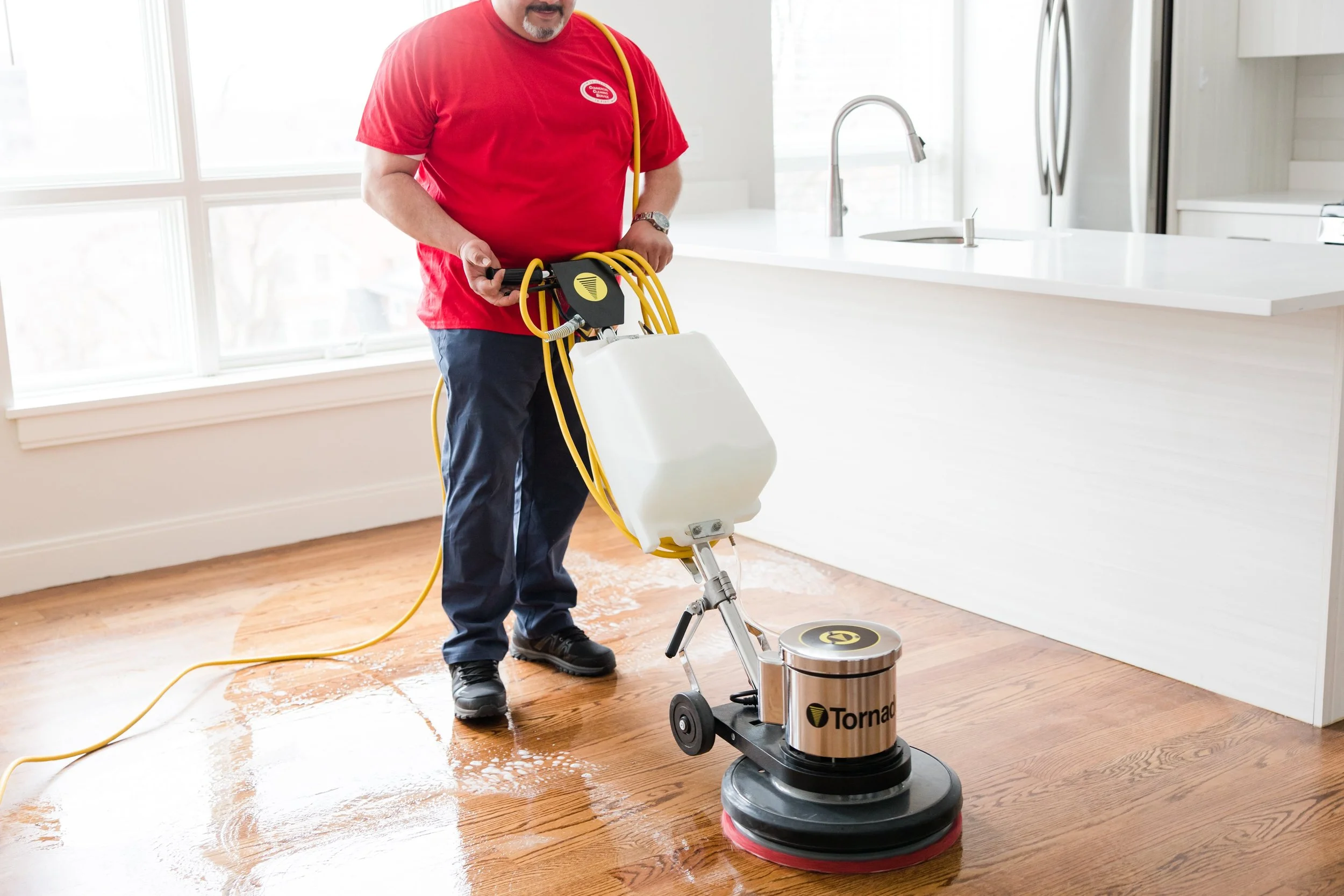 A man in a red shirt and dark pants is using a floor polishing machine on a hardwood floor, with soap suds around the machine in a bright kitchen.
