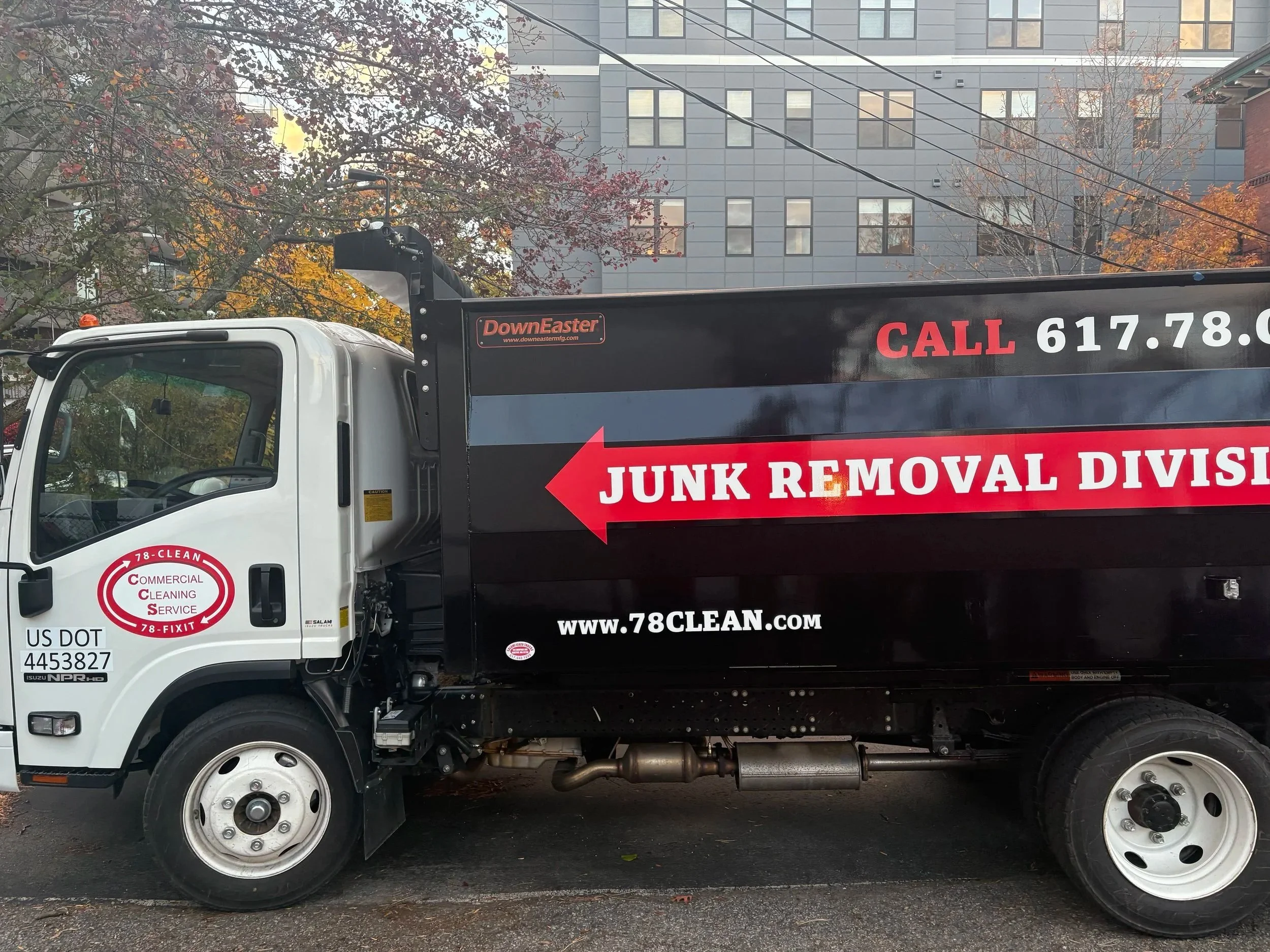 A white and black recycling truck parked on a street, with a background of buildings and trees with autumn foliage. The truck advertises junk removal services with a red arrow and white text on the black portion of the truck.