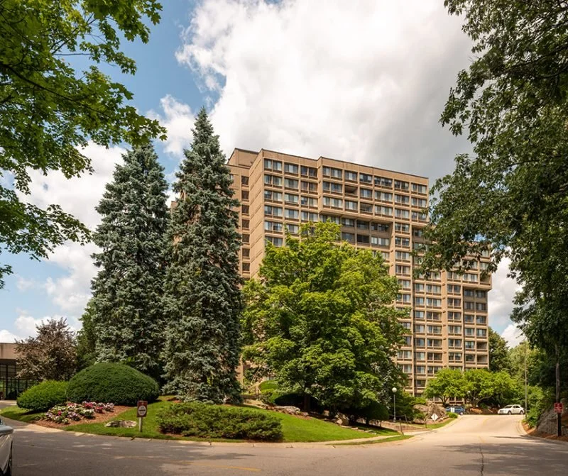 A multi-story apartment or office building surrounded by large trees and landscaped greenery, with a cloudy sky overhead.