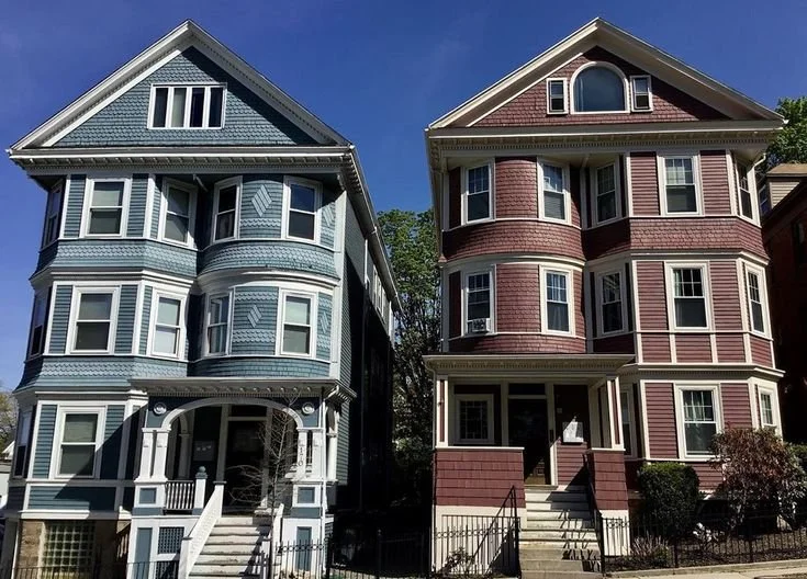 Two adjacent Victorian-style houses, one painted blue with white trim and the other painted red with white trim, under a clear blue sky.