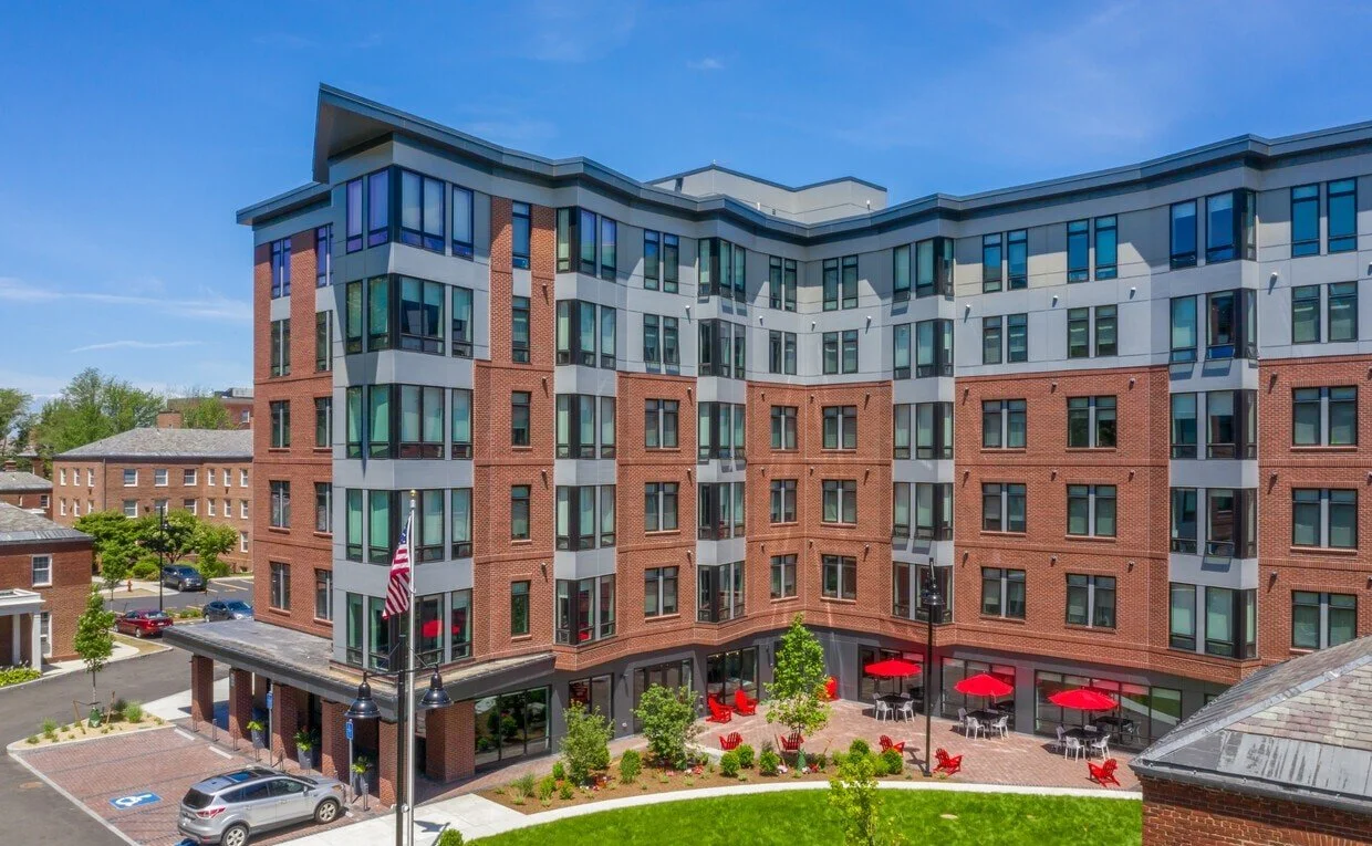 Modern multi-story residential building with red brick and gray exterior, large windows, outdoor seating area with red umbrellas, green landscaped yard, and parking lot, under a blue sky.