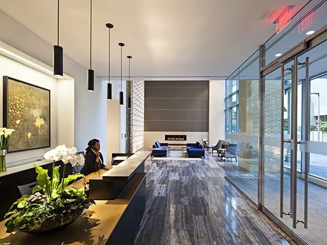 Modern hotel lobby with a reception desk, a woman working, and a seating area with blue and gray chairs, large windows, and hanging pendant lights.
