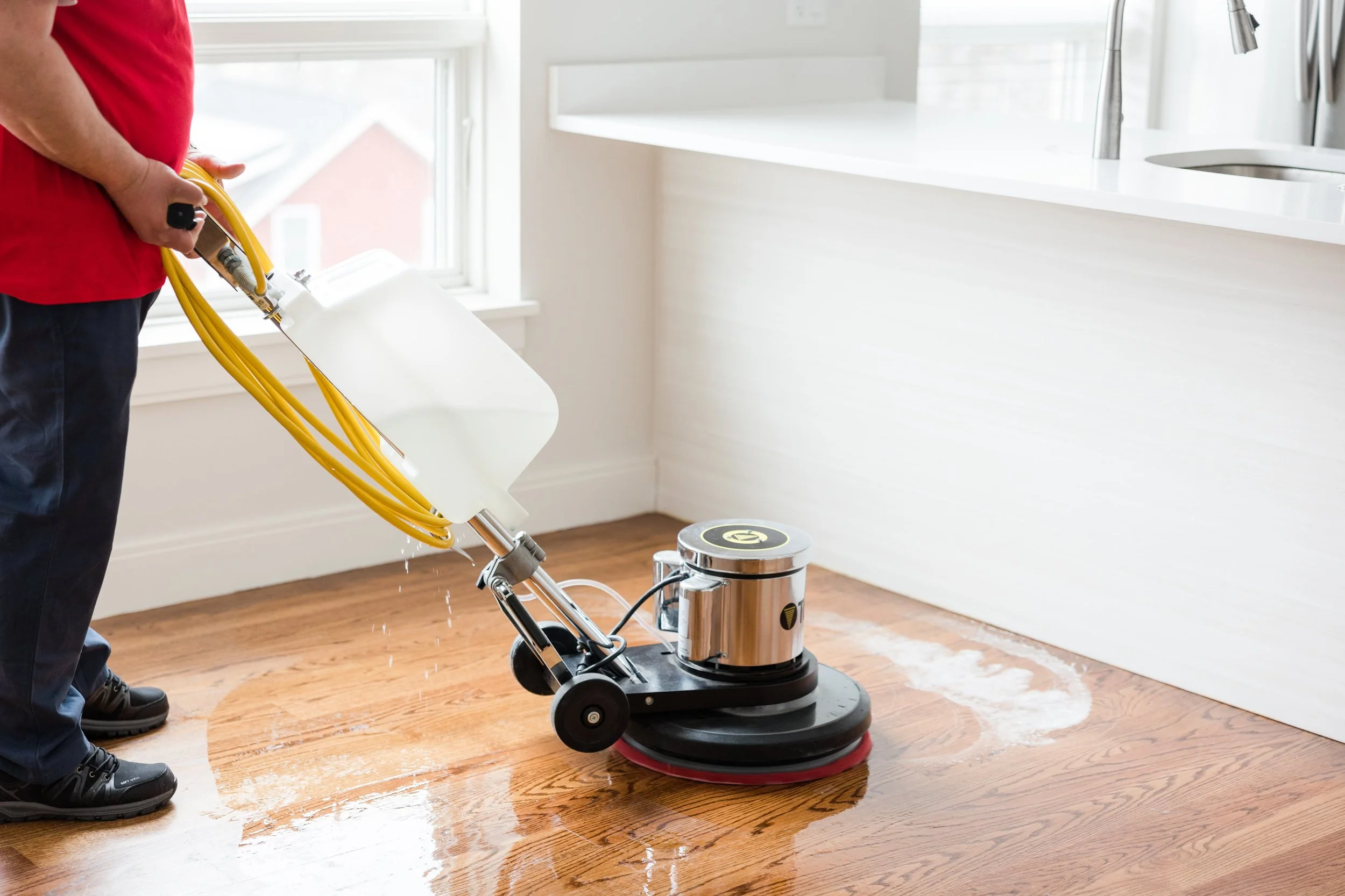 Person operating floor cleaning machine on hardwood floor in a bright kitchen.