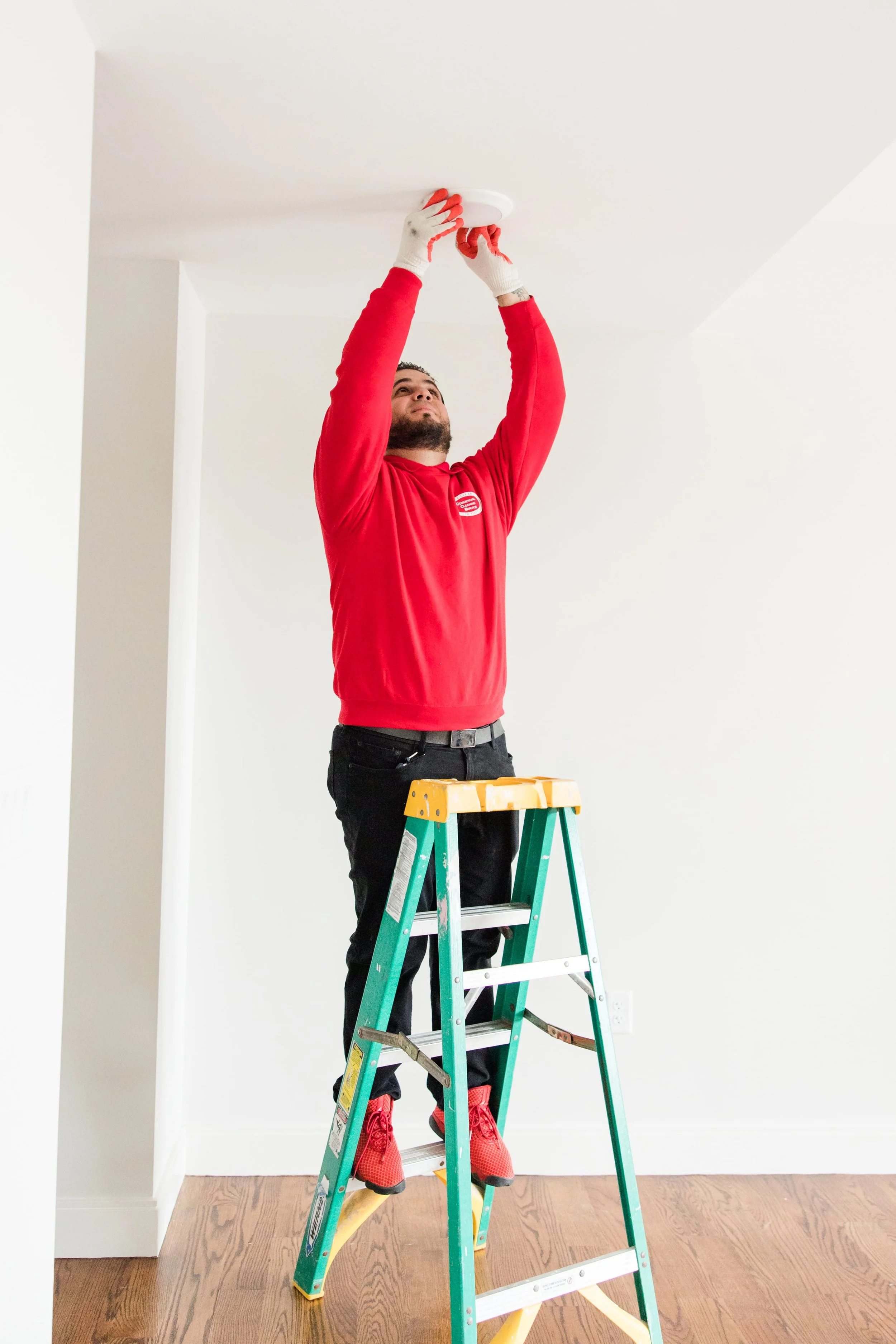 A man standing on a step ladder installing a smoke detector on a white ceiling in a room with white walls and wooden floor.