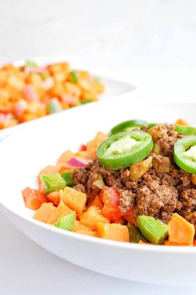 Close up of a white bowl filled with a sweet potato base and red onion, red pepper and green pepper topped with taco meat and sliced jalapenos