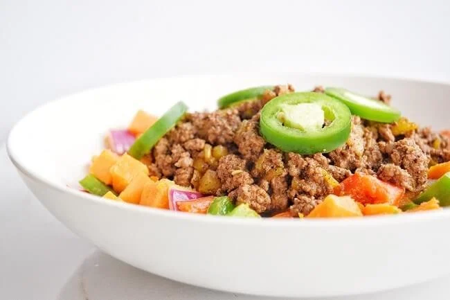 Side view of a bowl filled with a sweet potato, red onion, red pepper and green pepper base topped with taco meat and sliced jalapenos