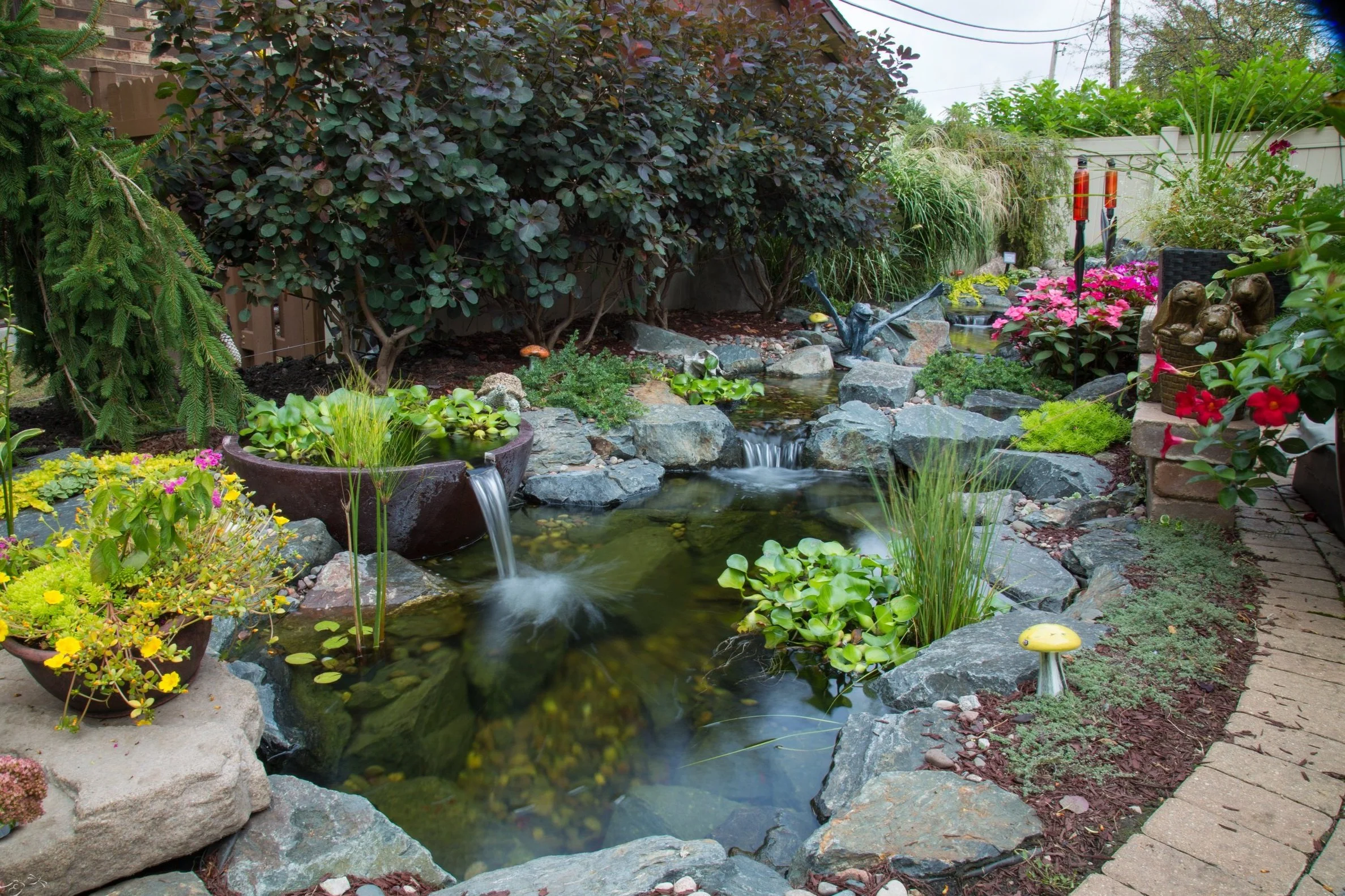 A backyard garden with a stream flowing over rocks, surrounded by colorful flowers and green plants, with a patio on the right side and bushes in the background.