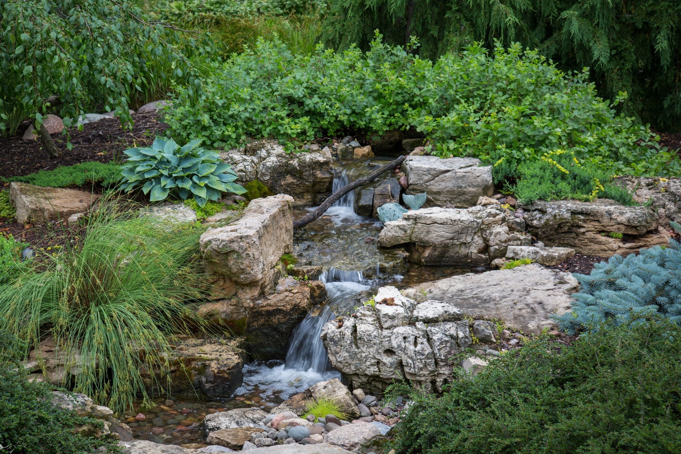 Small waterfall flowing over rocks surrounded by lush green plants in a garden.