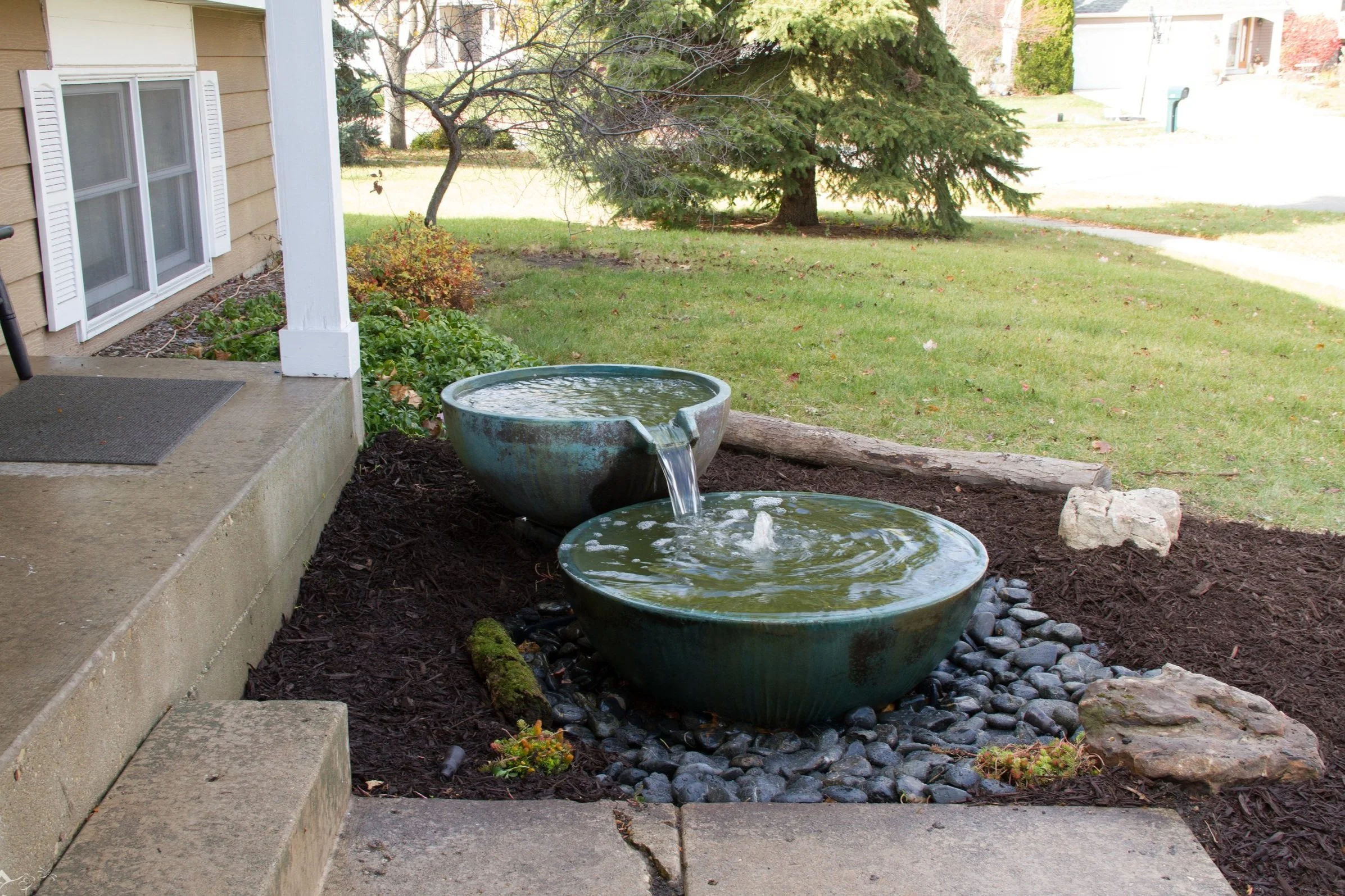 Outdoor scene with two ceramic water bowls on black rocks in front of a house, with a green lawn, trees, and a driveway in the background.