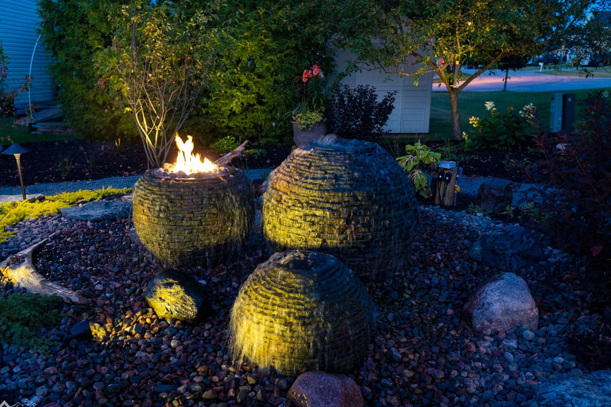 A backyard garden at dusk featuring a water fountain with three tiered bowls, illuminated by outdoor lighting, surrounded by rocks, plants, and trees.