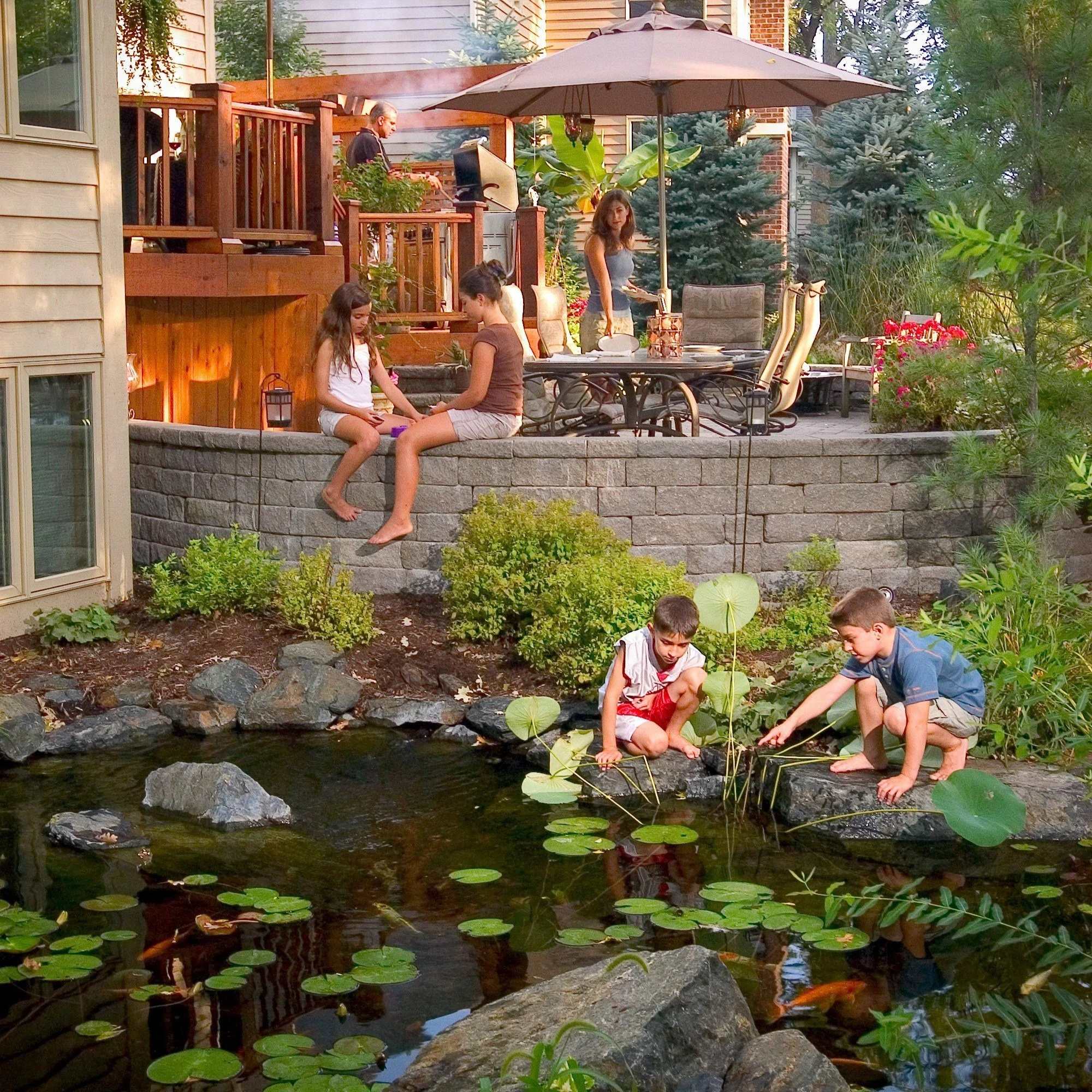 A backyard scene with children playing by a pond with water lilies, family members on a raised deck with outdoor furniture, a person grilling, and lush greenery around.