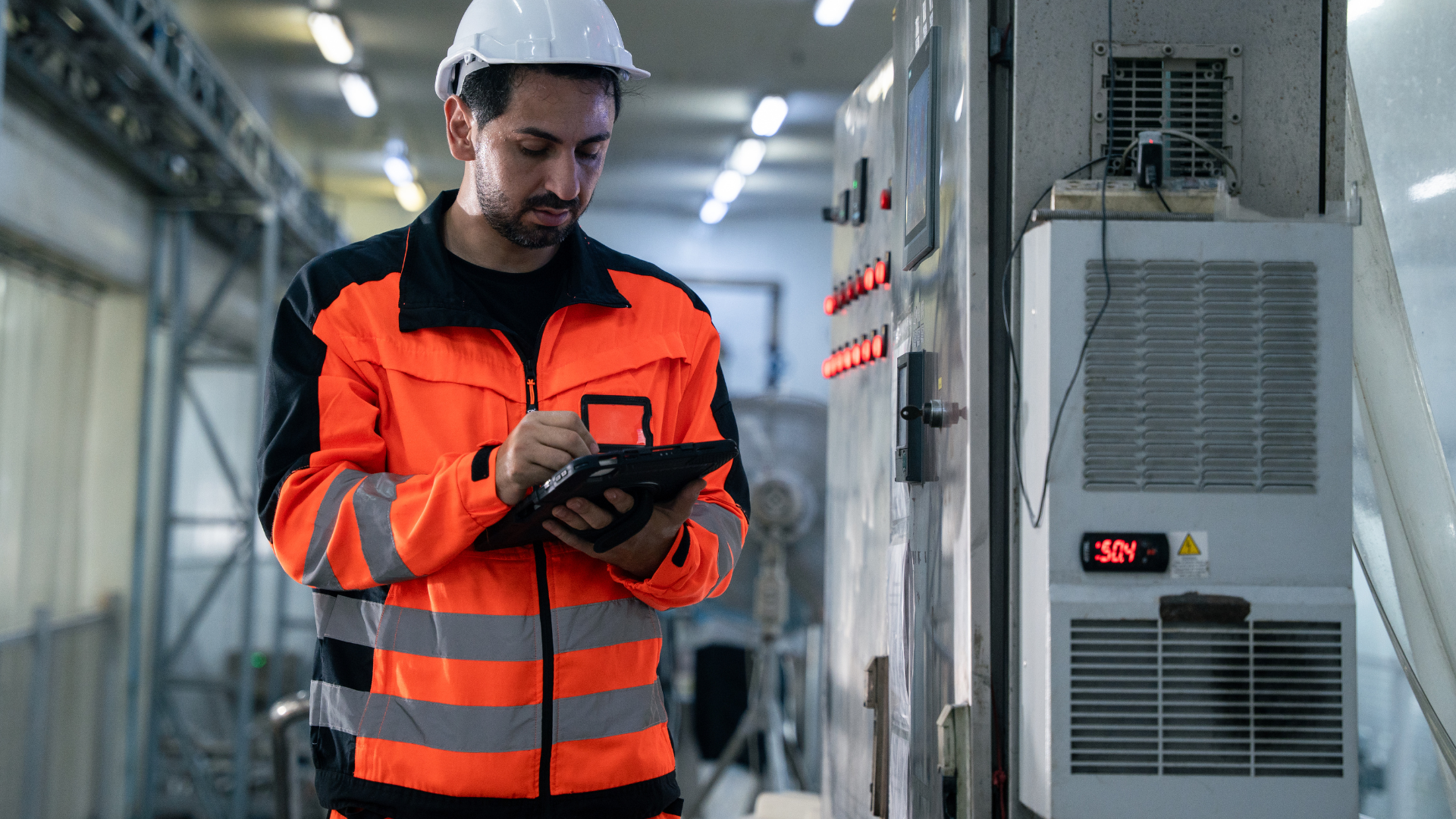 Operaio in fabbrica con giacca arancione e caschetto bianco, che utilizza un dispositivo digitale davanti a un macchinario industriale in un ambiente di produzione.