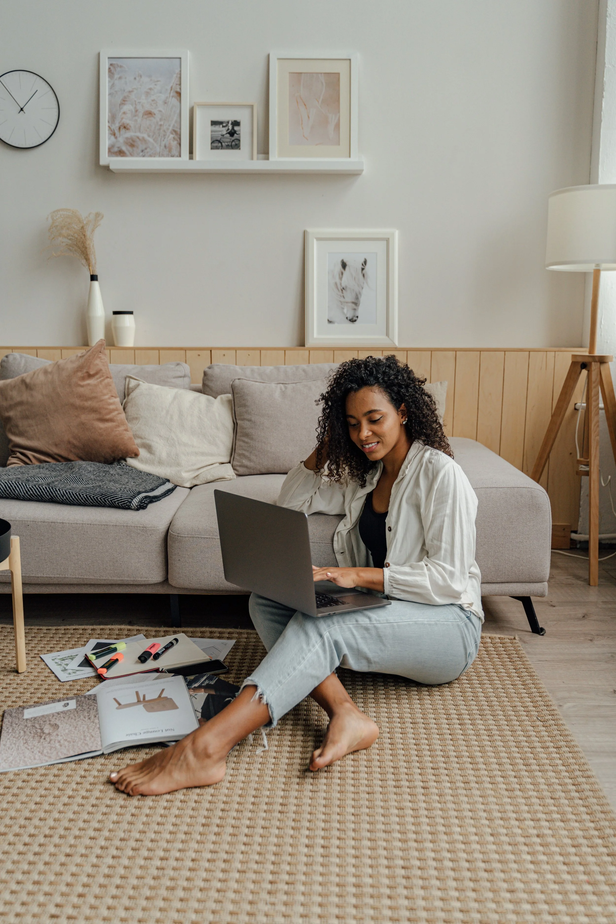 A woman sitting on a rug in a living room, working on a laptop, with open magazines, notebooks, and highlighters scattered around.