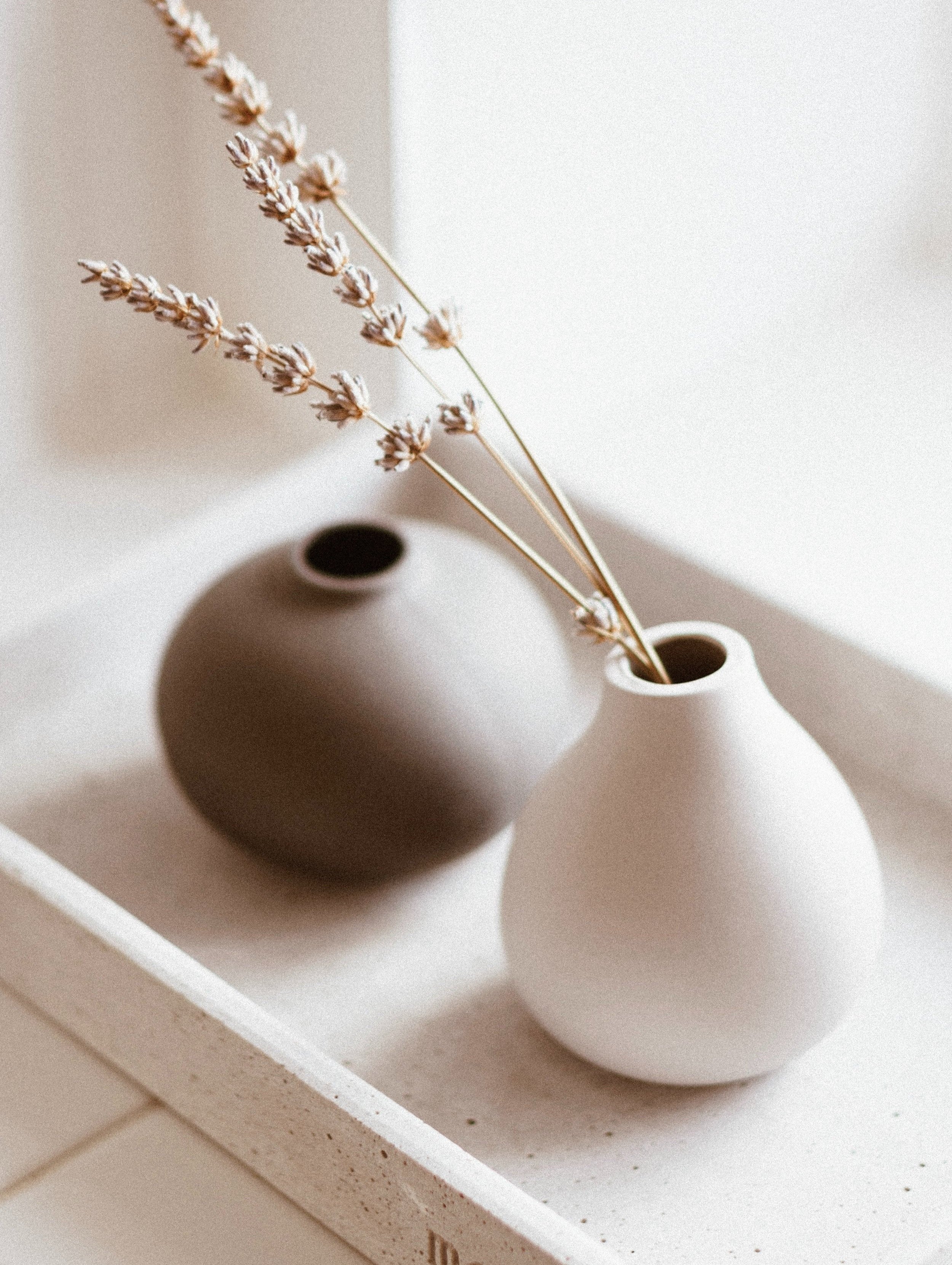 Two minimalist ceramic vases, one light-colored and one dark-colored, on a white surface with dried lavender in the light-colored vase.