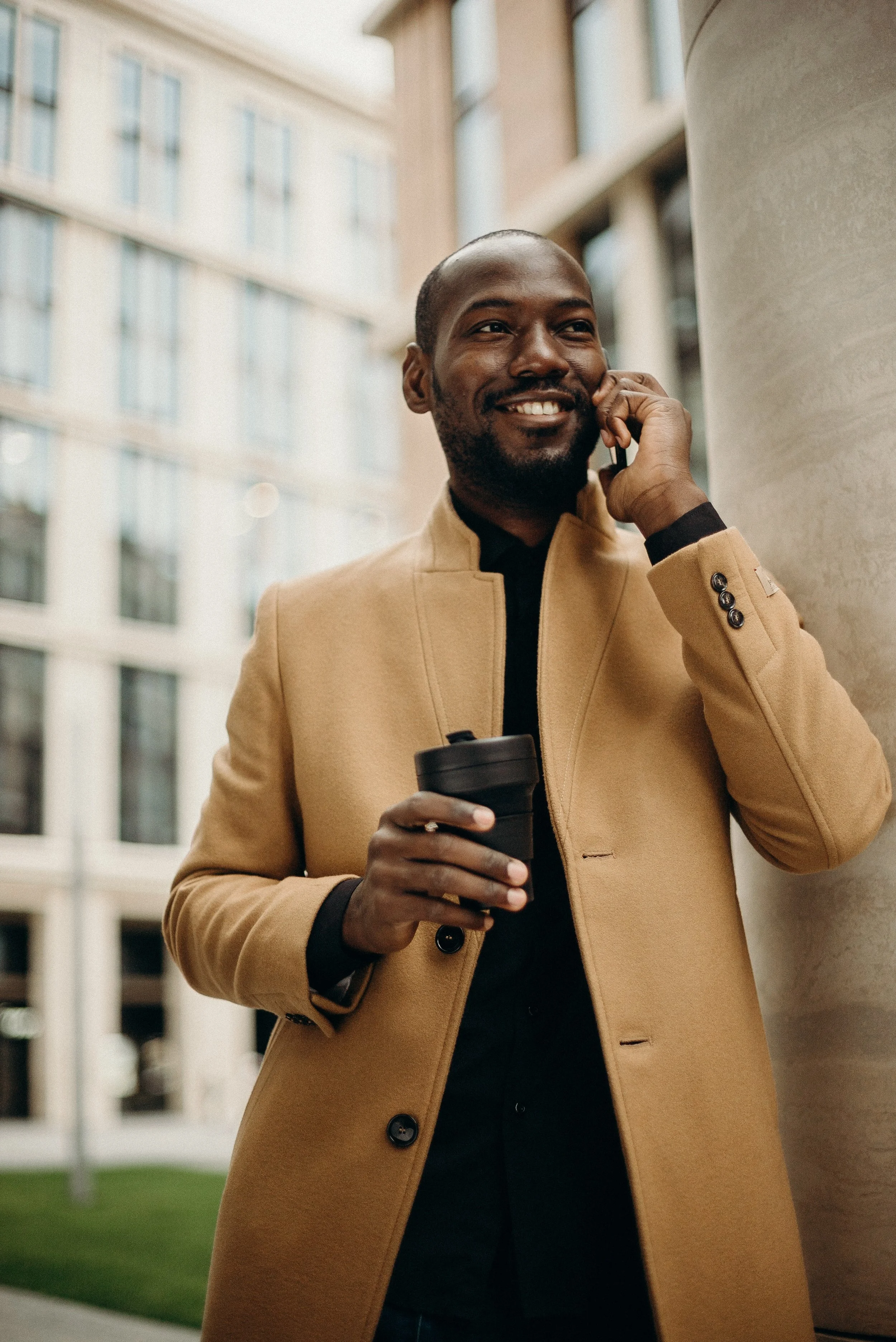 A man dressed in a tan coat talking on a cellphone while holding a coffee cup, standing outdoors near a column with modern office buildings in the background.