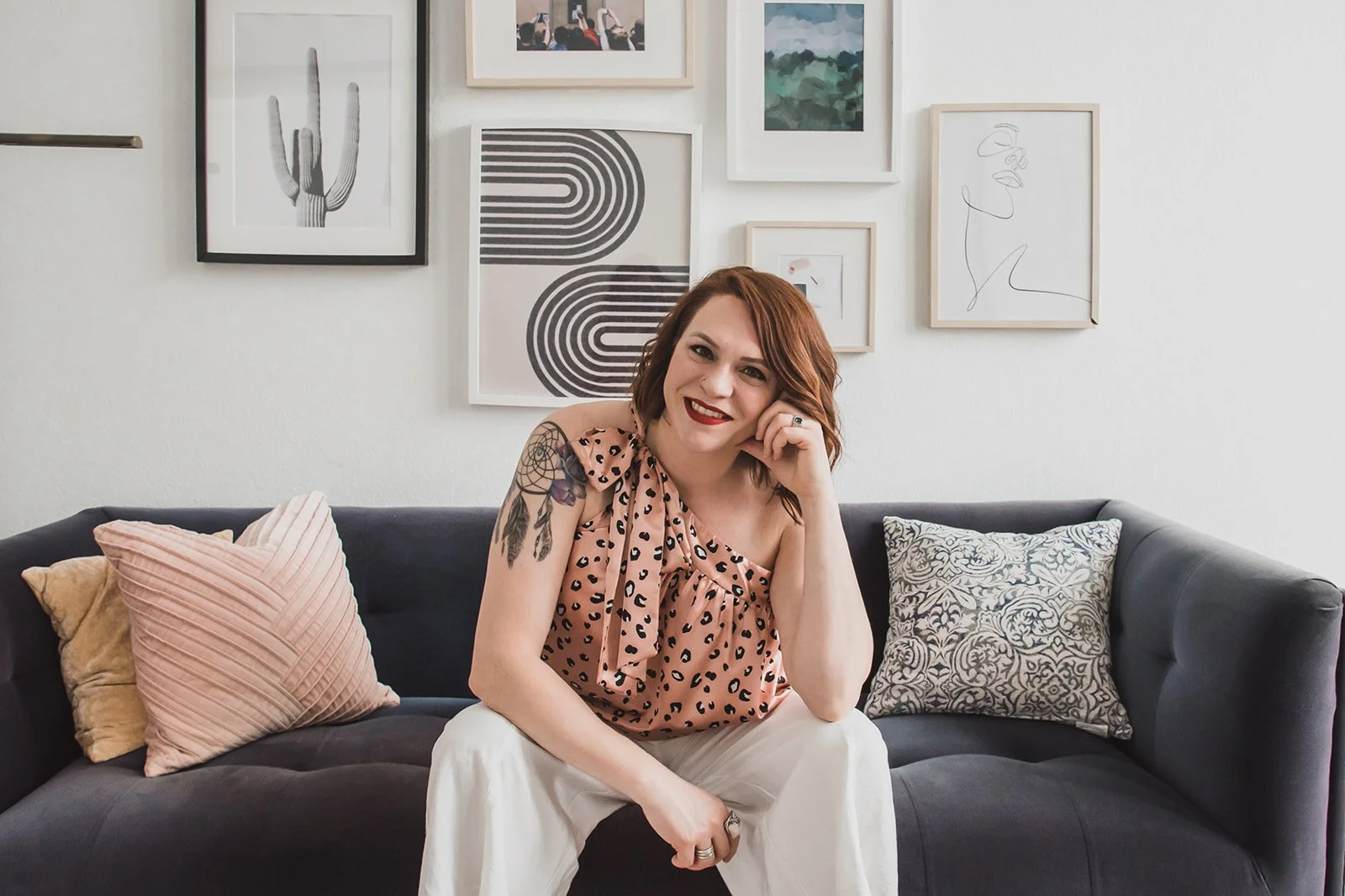 A young woman with red hair and tattoos on her left arm, sitting on a black sofa, smiling, with her right hand resting on her chin. She is wearing a sleeveless, leopard print top and white pants. Behind her, there is a white wall decorated with various framed art pieces, including a cactus, abstract patterns, and line art.
