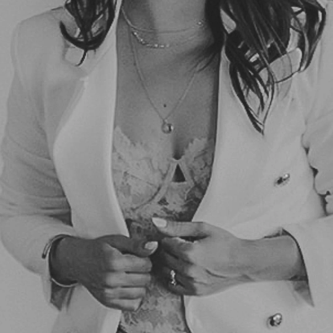 Close-up of a person wearing a white blazer over a lace top, with visible necklaces and ringed fingers.