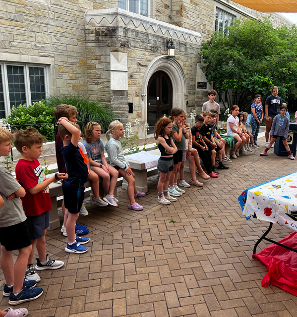 Children sitting on a ledge outside a stone building, some are standing and some are seated, participating in an outdoor activity or game on a brick patio.