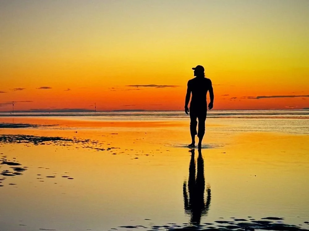 A lone figure on a wet beach at sunset, reflected in the sand
