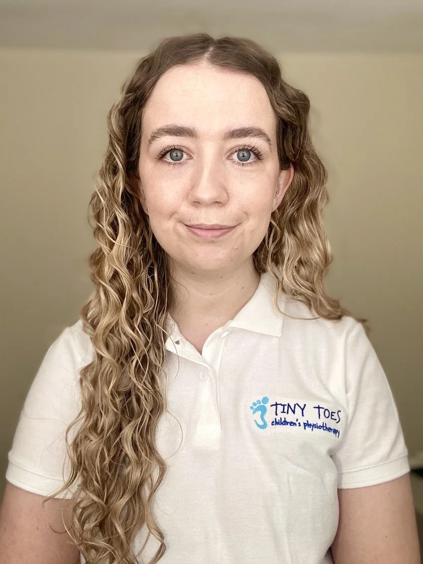 A woman with long, curly blonde hair wearing a white polo shirt with a logo that says "Tiny Toes Children's Physiotherapy".