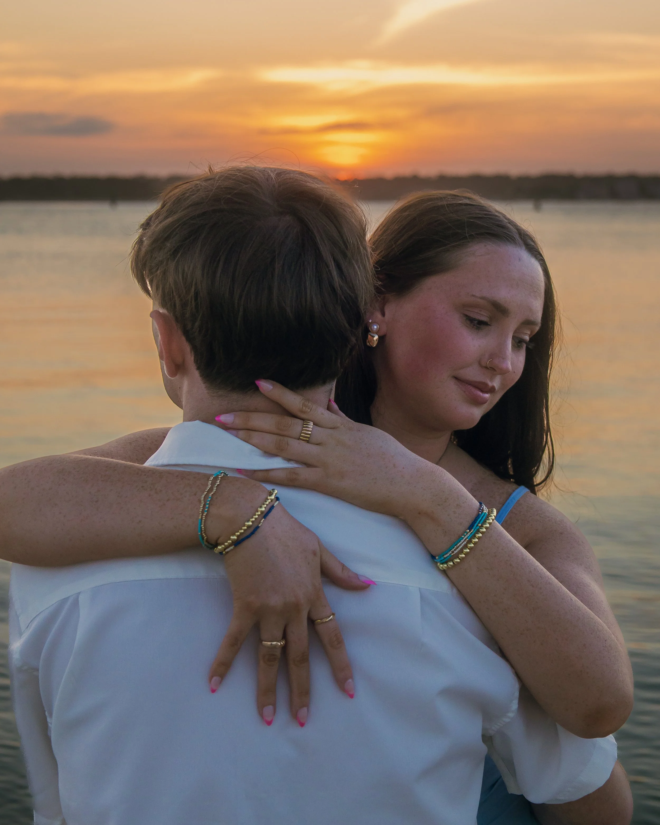 Two women embracing at sunset by a body of water, with the focus on their faces and jewelry, and the sunset illuminating the scene.