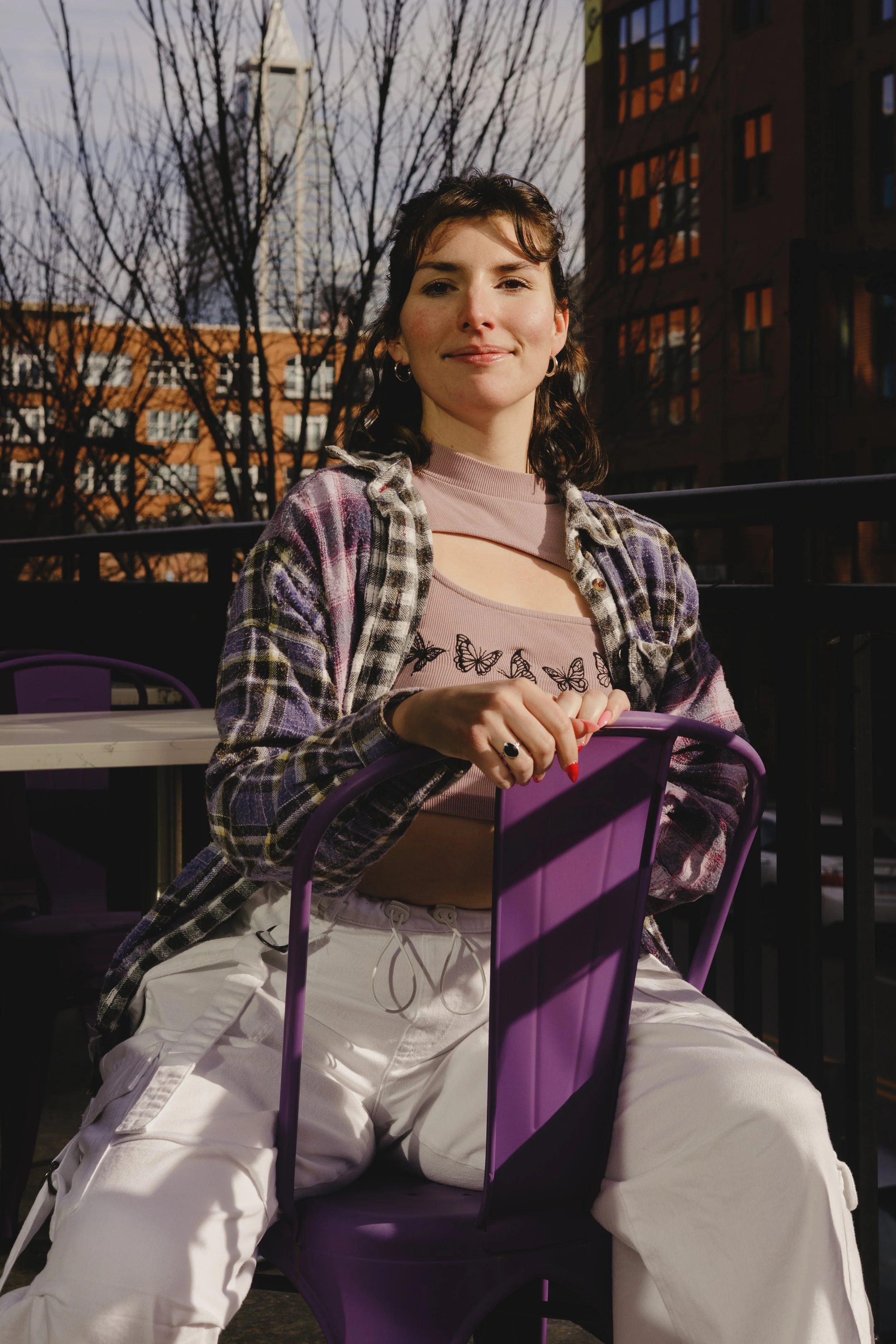 A young woman with dark brown hair and earrings smiling while sitting at an outdoor cafe table during evening or dusk, with leafless trees and tall buildings in the background.