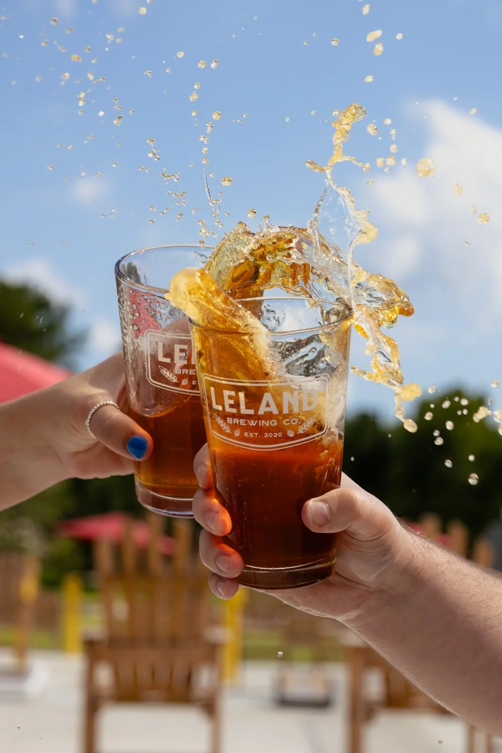Three people holding glasses of beer in a toast outdoors, with splashing beer and a clear blue sky.