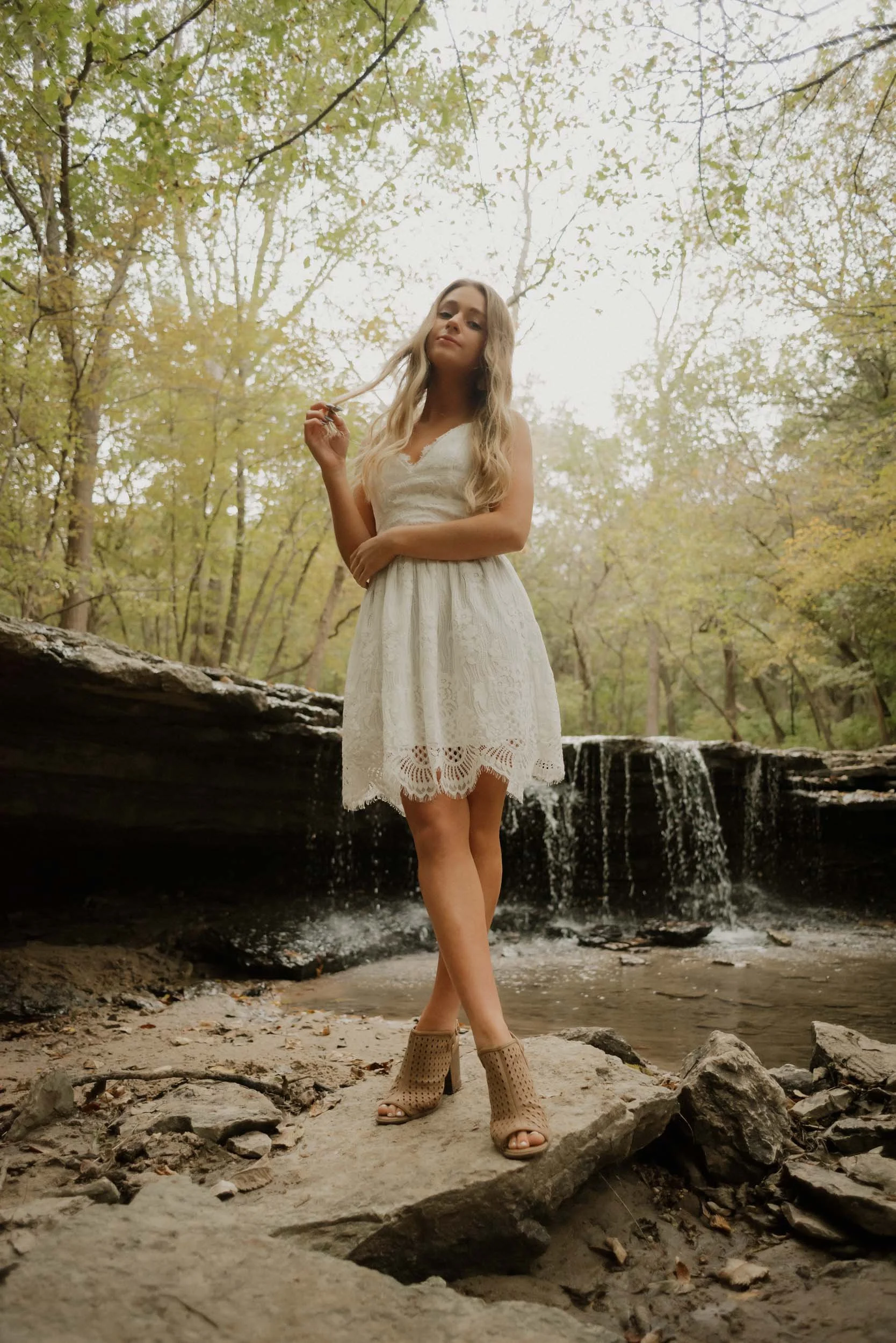 A woman in a white lace dress and tan open-toe heels stands on a large rock near a stream with small waterfalls in a wooded area.