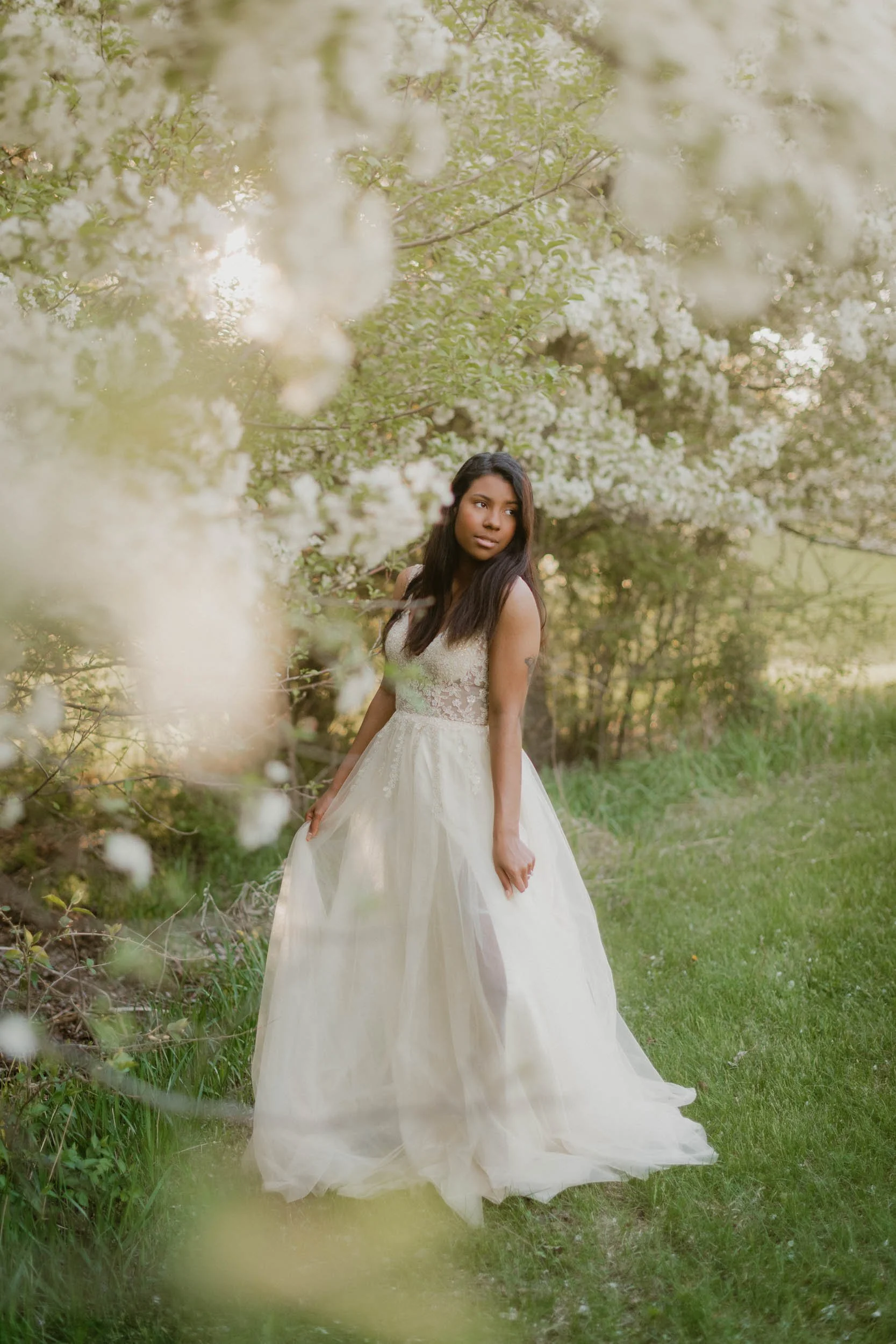 A woman in a white gown standing outdoors among blooming trees with sunlight filtering through the branches.