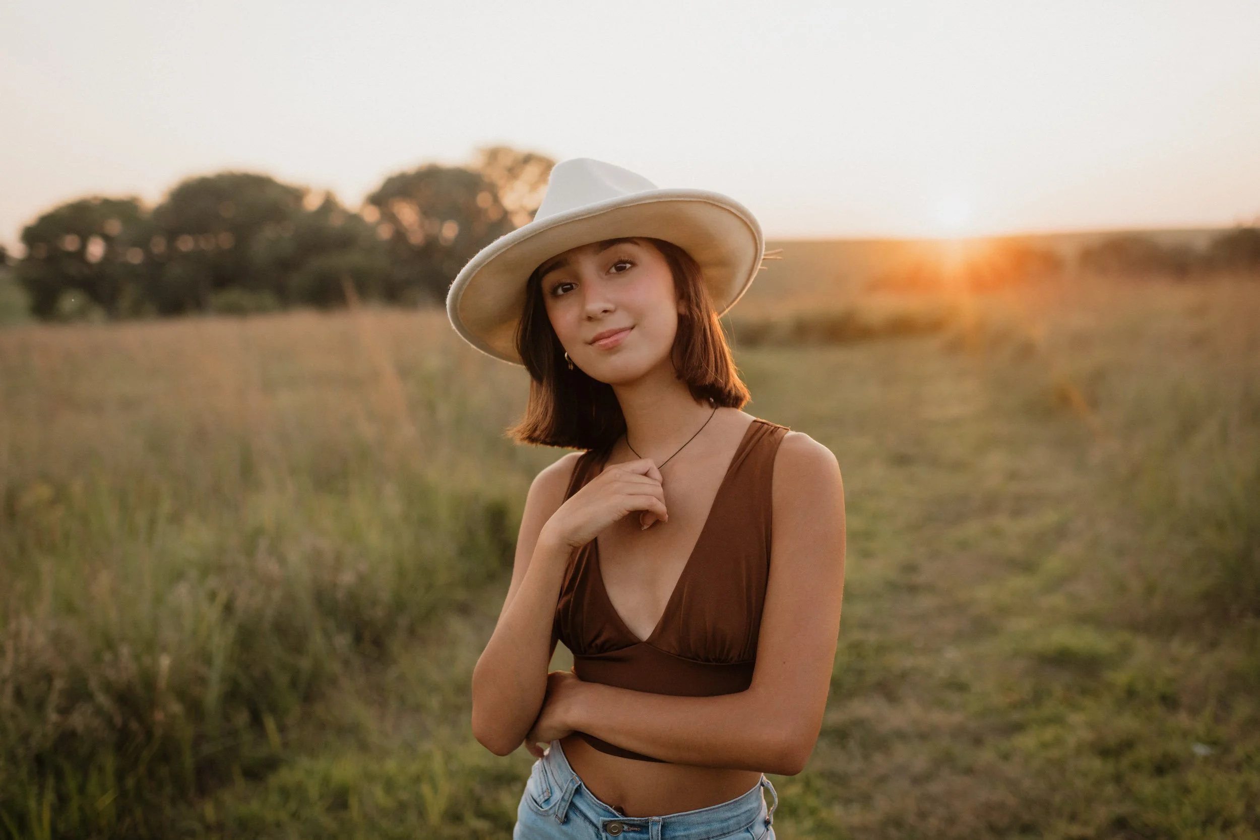 A Nebraska senior portrait of a young woman wearing a white wide-brimmed hat and a brown sleeveless top standing in a field at sunset.
