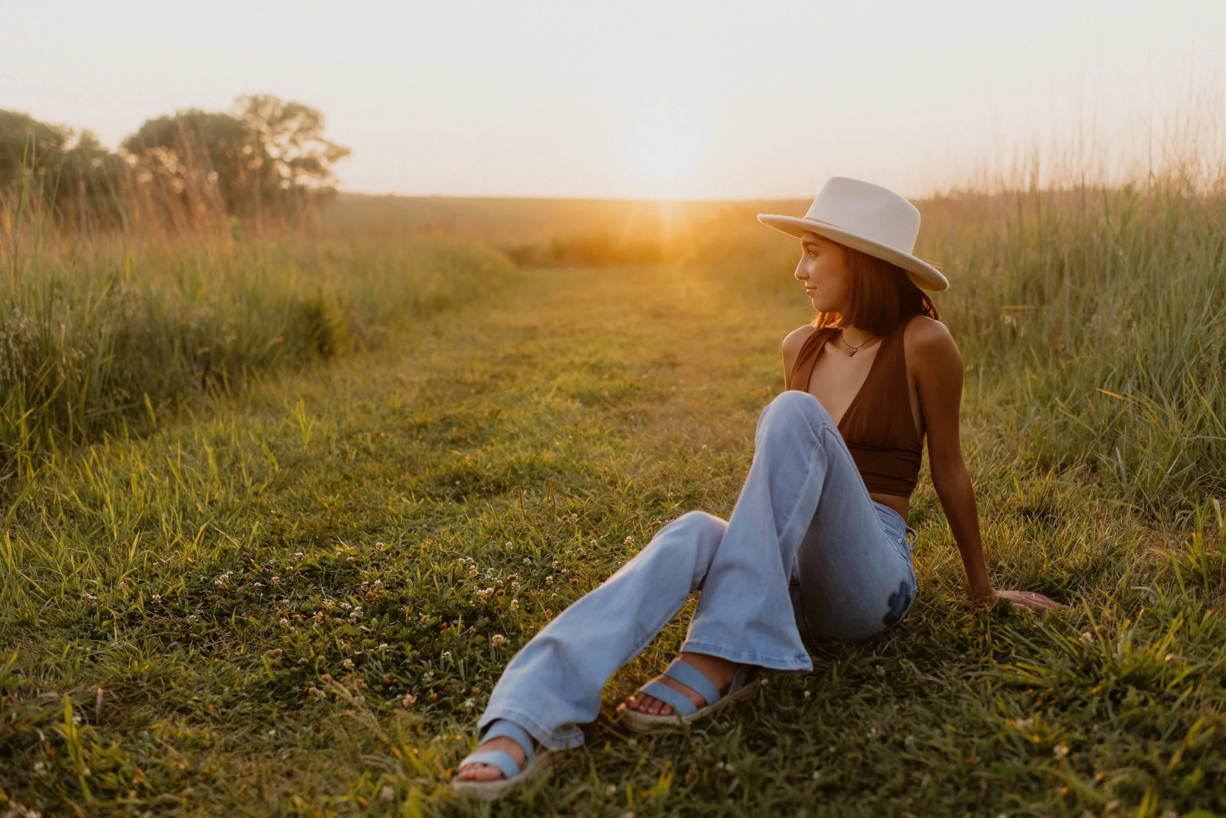 A young woman sitting on the grass in a field during sunset in Lincoln Nebrakska, wearing a wide-brimmed hat, brown top, light jeans, and sandals. 