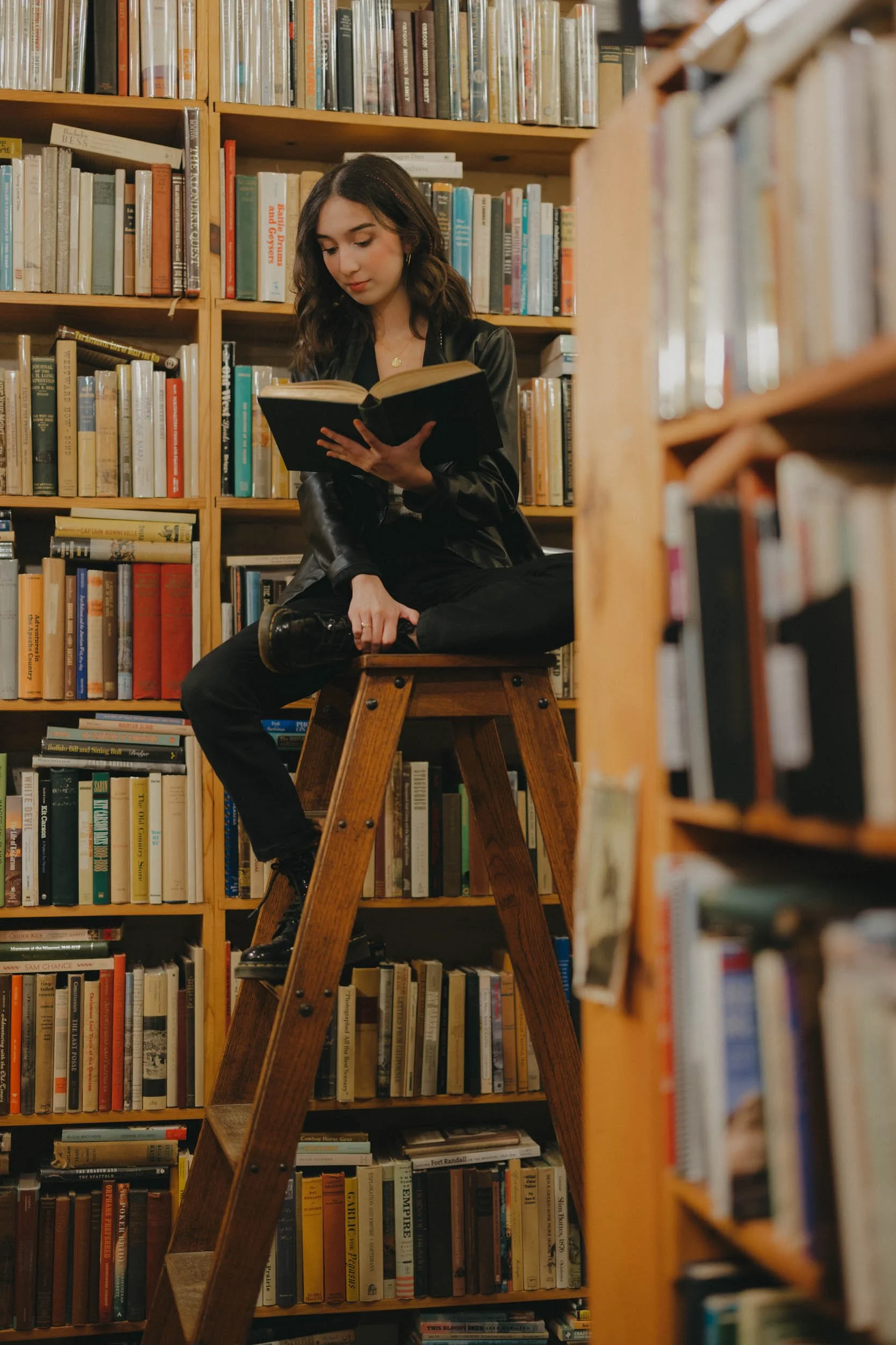 A young woman sitting on a wooden ladder in a library, reading a book, surrounded by bookshelves filled with books