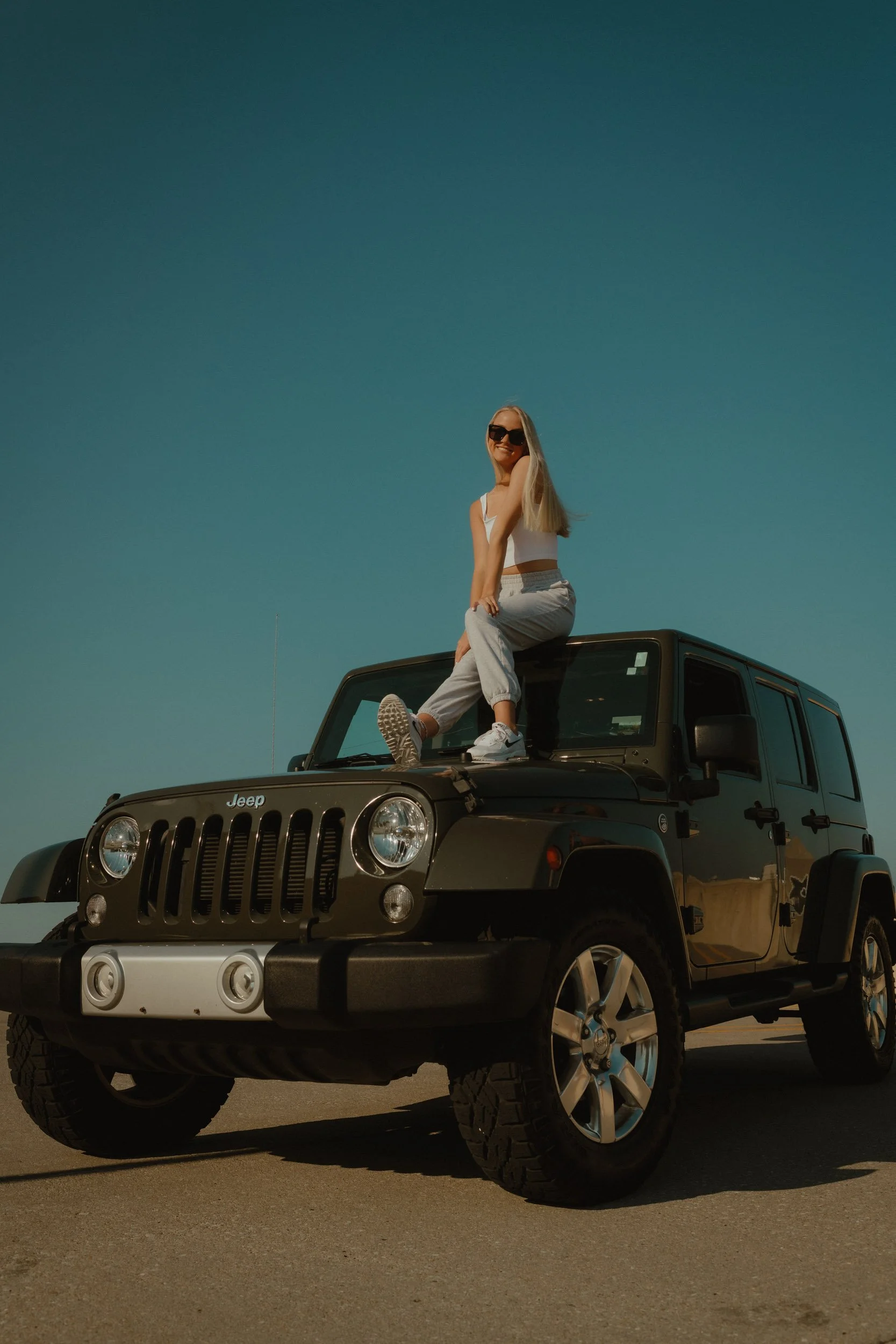 A woman with long blonde hair wearing sunglasses, a white tank top, and light-colored pants sitting on the hood of a black Jeep Wrangler, smiling with a clear sky in the background.