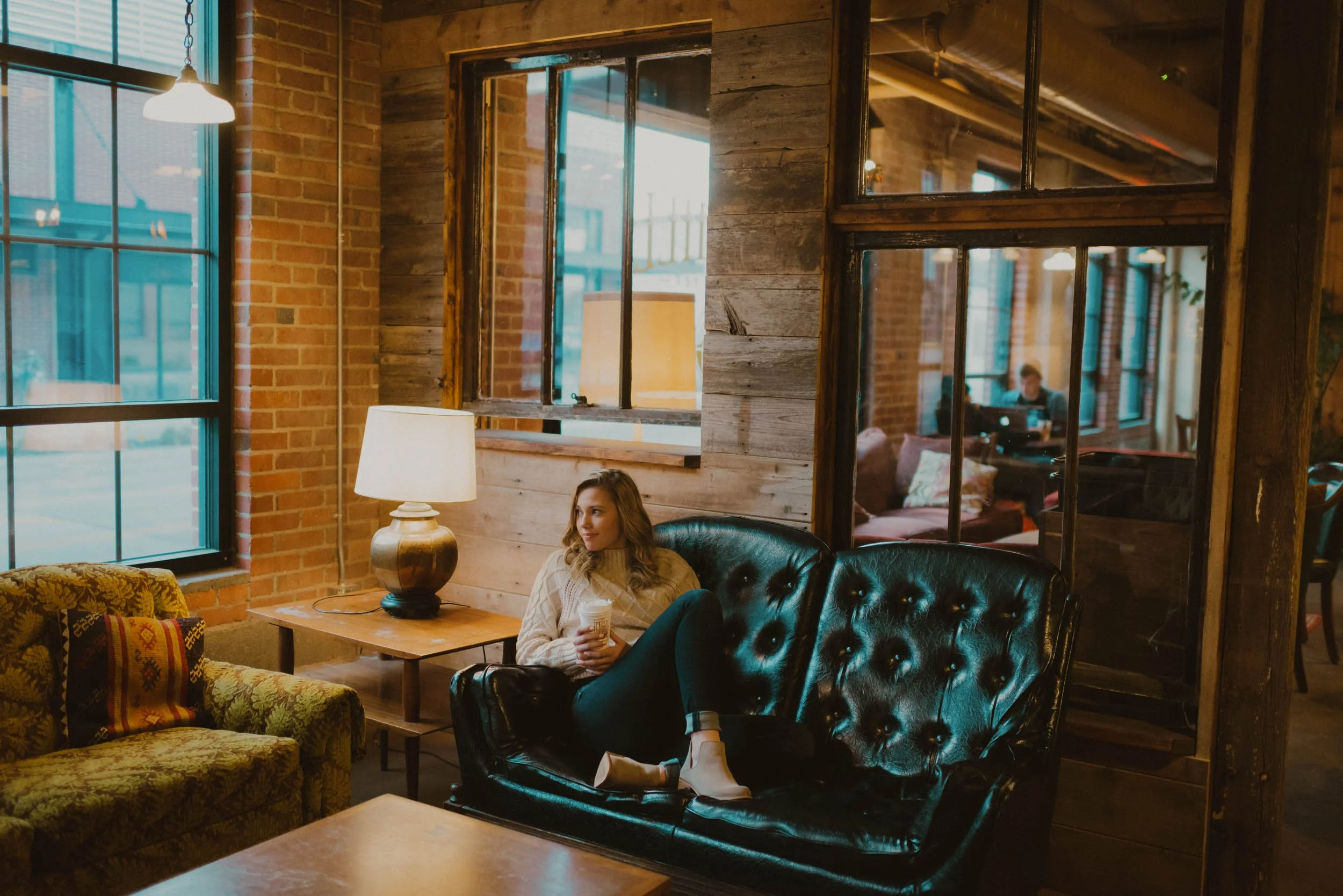 A woman sitting on a black leather couch inside a cozy, rustic-style coffee shop, holding a coffee mug, with warm lighting, exposed brick walls, large windows, and a person working on a laptop in the background.