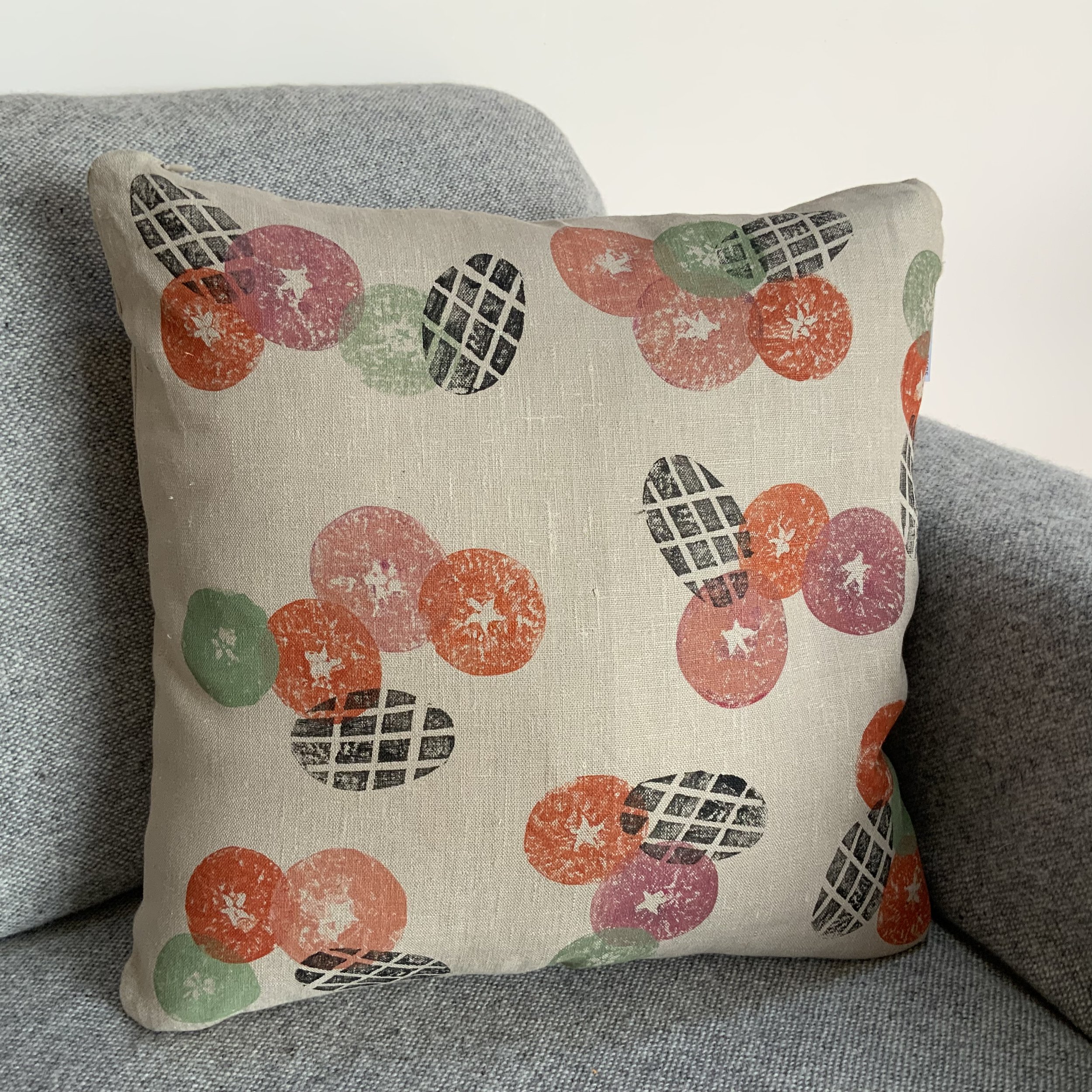 Hand-printed cushionin Devon by Susie Honnor using apples potatoes on 100%natural linen. A rich orange and red contrasts with a fresh green carefully over-printed with black. Viewed on sofa.