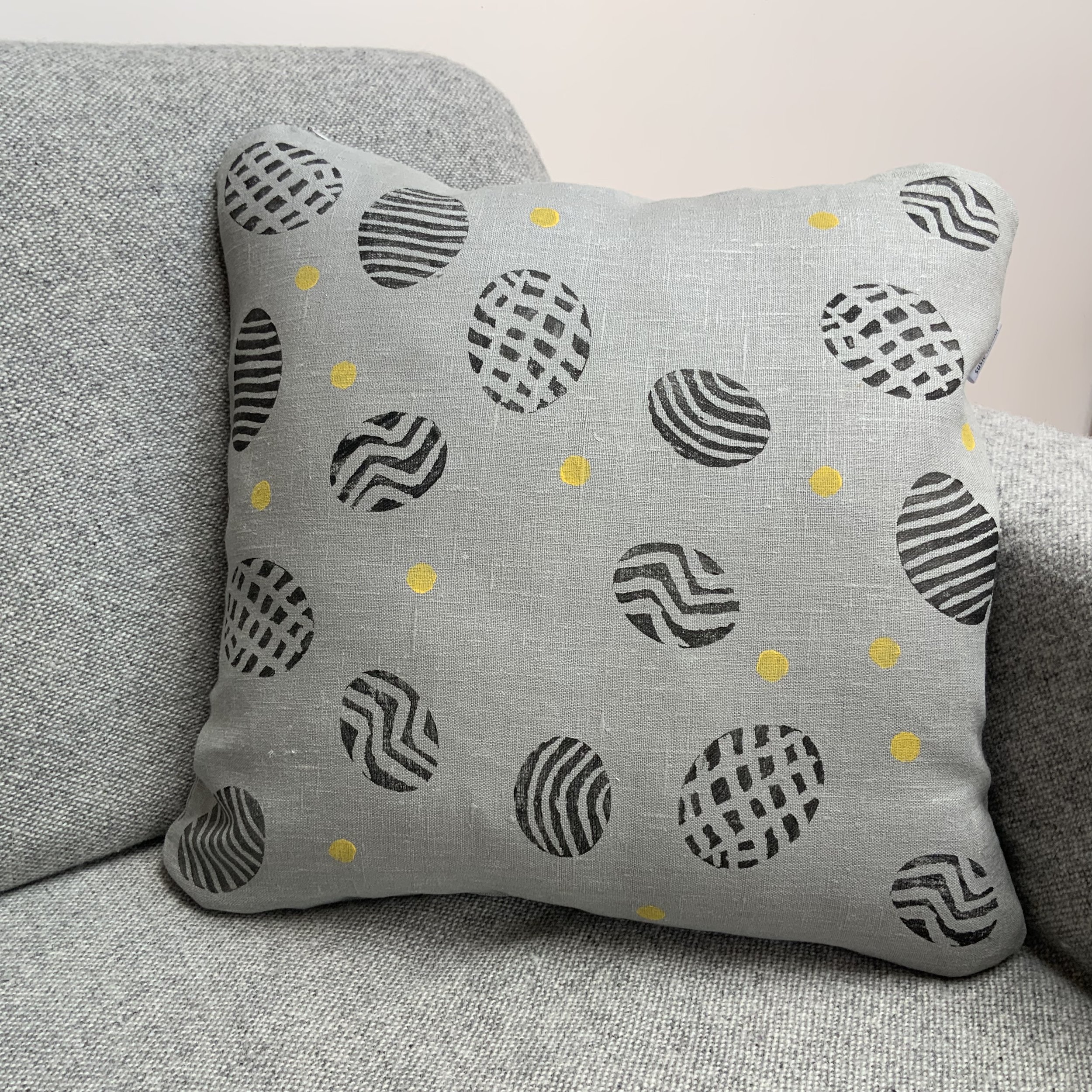 Potato print in black patterns with yellow spots on 100%grey linen. Cushio sits on a sofa.