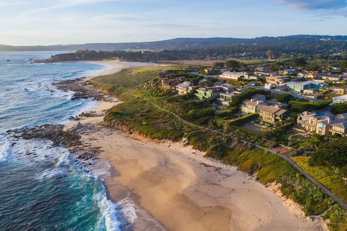 Aerial view of a coastal residential neighborhood with houses near a sandy beach and rocky shoreline, with hills and trees in the background.