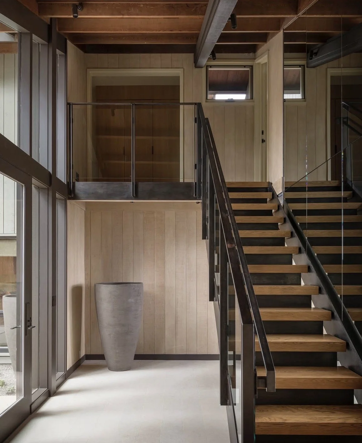 Interior view of a modern staircase with wooden steps, black handrails, and a glass railing, in a room with wood-paneled walls and a large gray vase on the floor near glass doors.