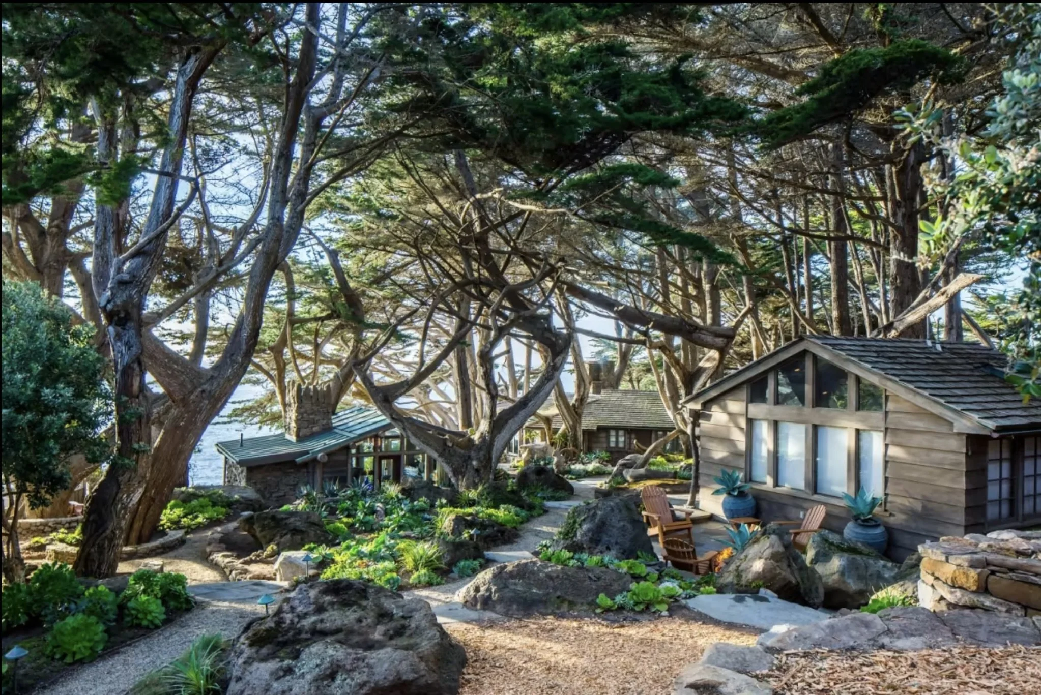 Cozy outdoor garden scene with trees, rocks, and small wooden cottages, featuring seating areas and potted plants.