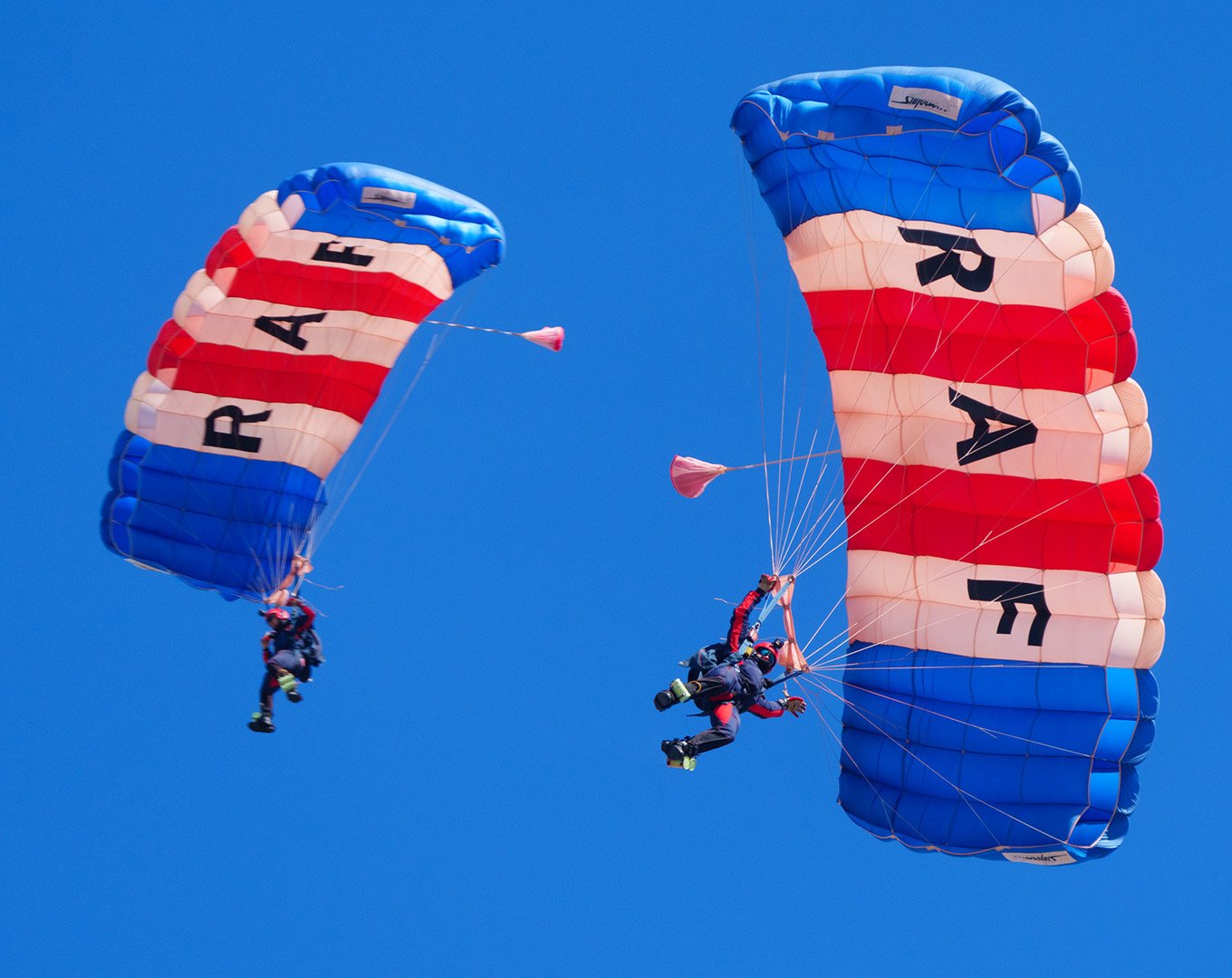 Two skydivers descend with parachutes decorated with red, white, and blue stripes and the word "USA" on each chute, against a clear blue sky.