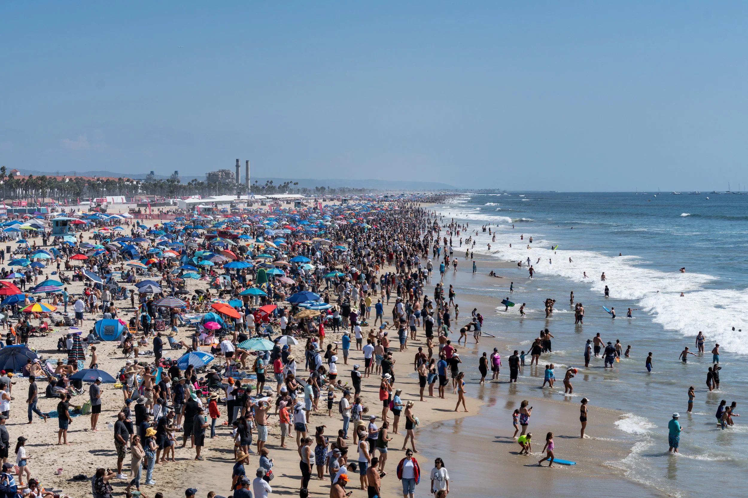 Crowded beach filled with umbrellas, beachgoers, and people swimming in the ocean under a clear blue sky.