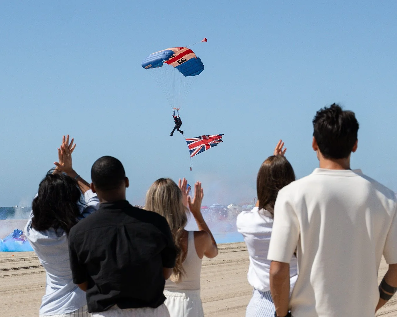 5 people clapping as a parachute jumper lands on the beach with a flag.,