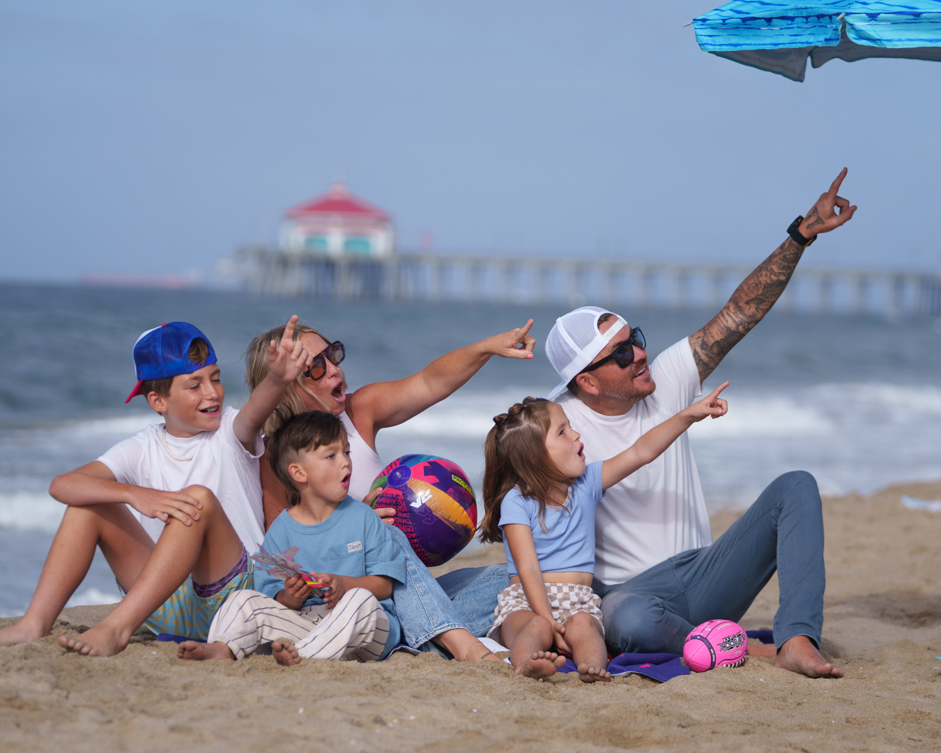 Family of 5 sitting on the beach in front of Huntington Beach Pier, pointing at the sky.