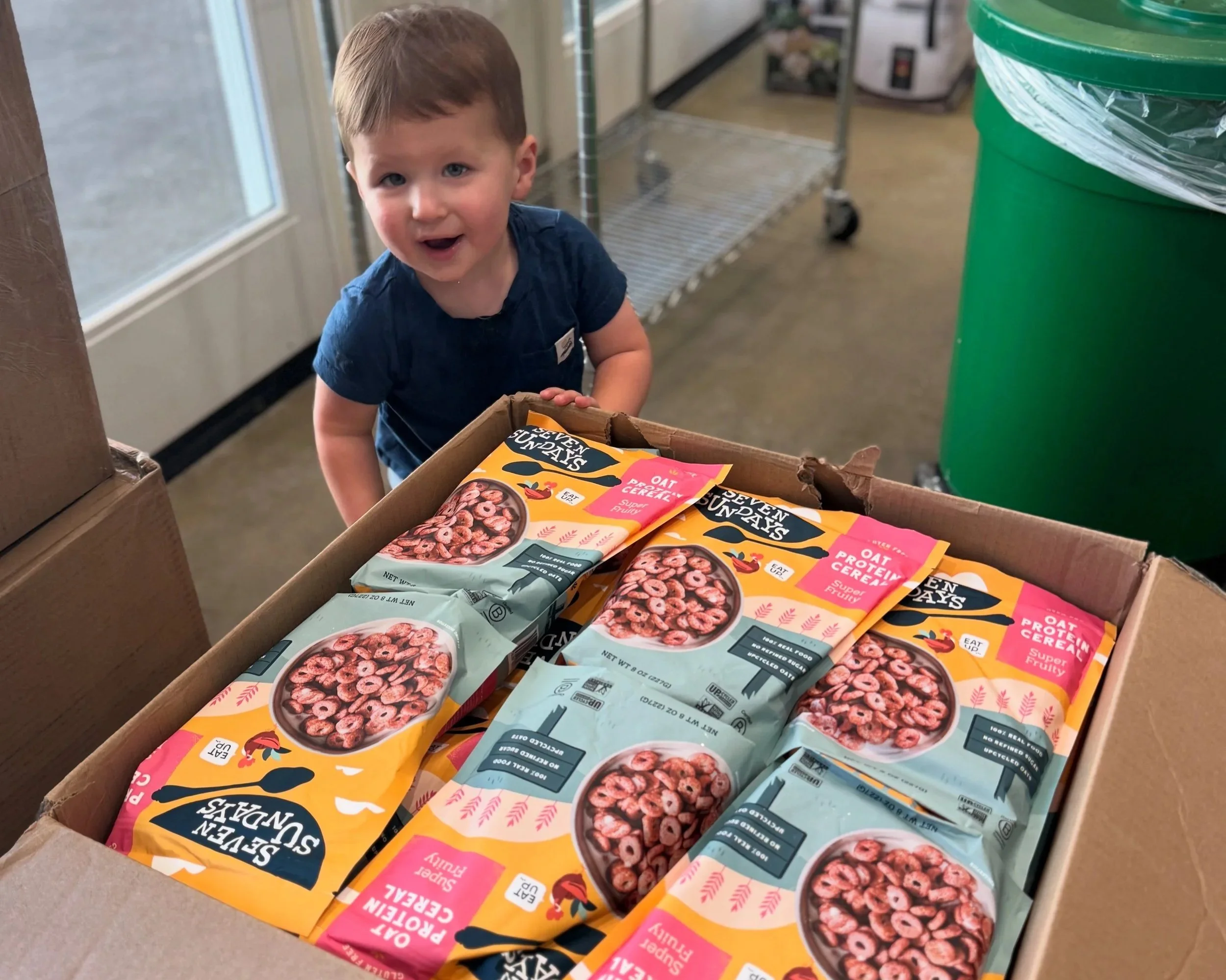A young girl is lying at a table with her head resting on her arm, watching an adult pour food into a small pink bowl. The girl is holding a pink candle with a white wick in her other hand, and there is a pink container beside her. The background shows a bright room with large windows and trees outside.