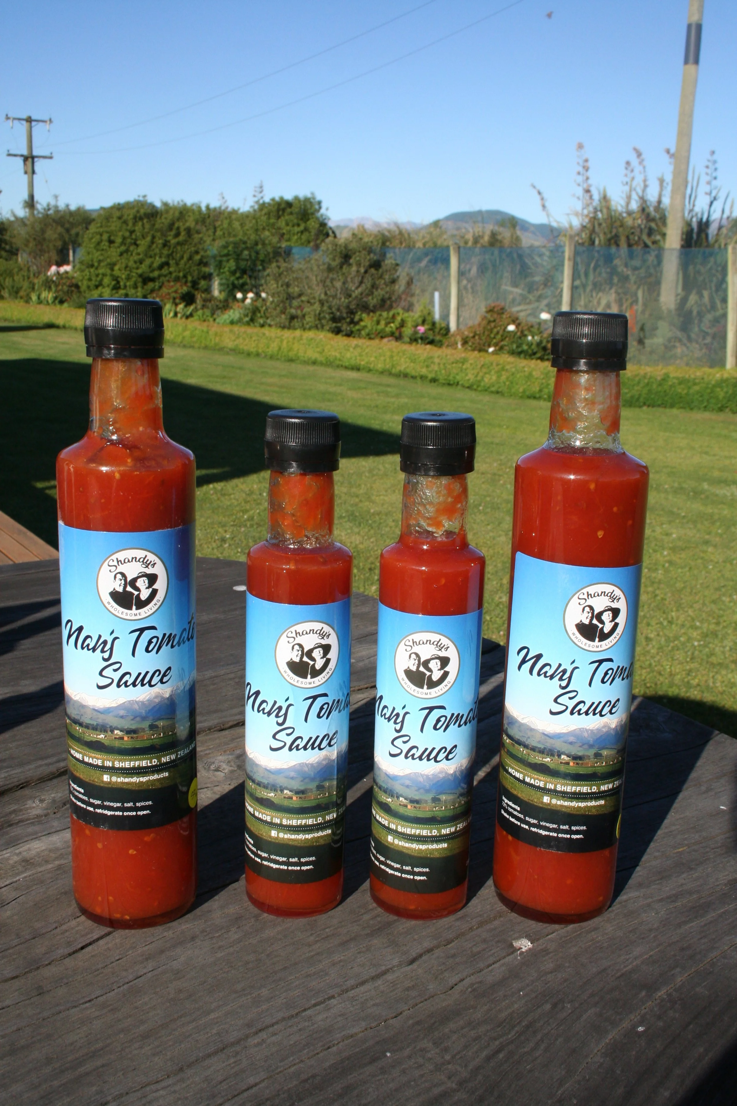 Four bottles of Shandy's Nan's Tomato Sauce on a wooden table outdoors, with a grassy yard, trees, mountains in the background.