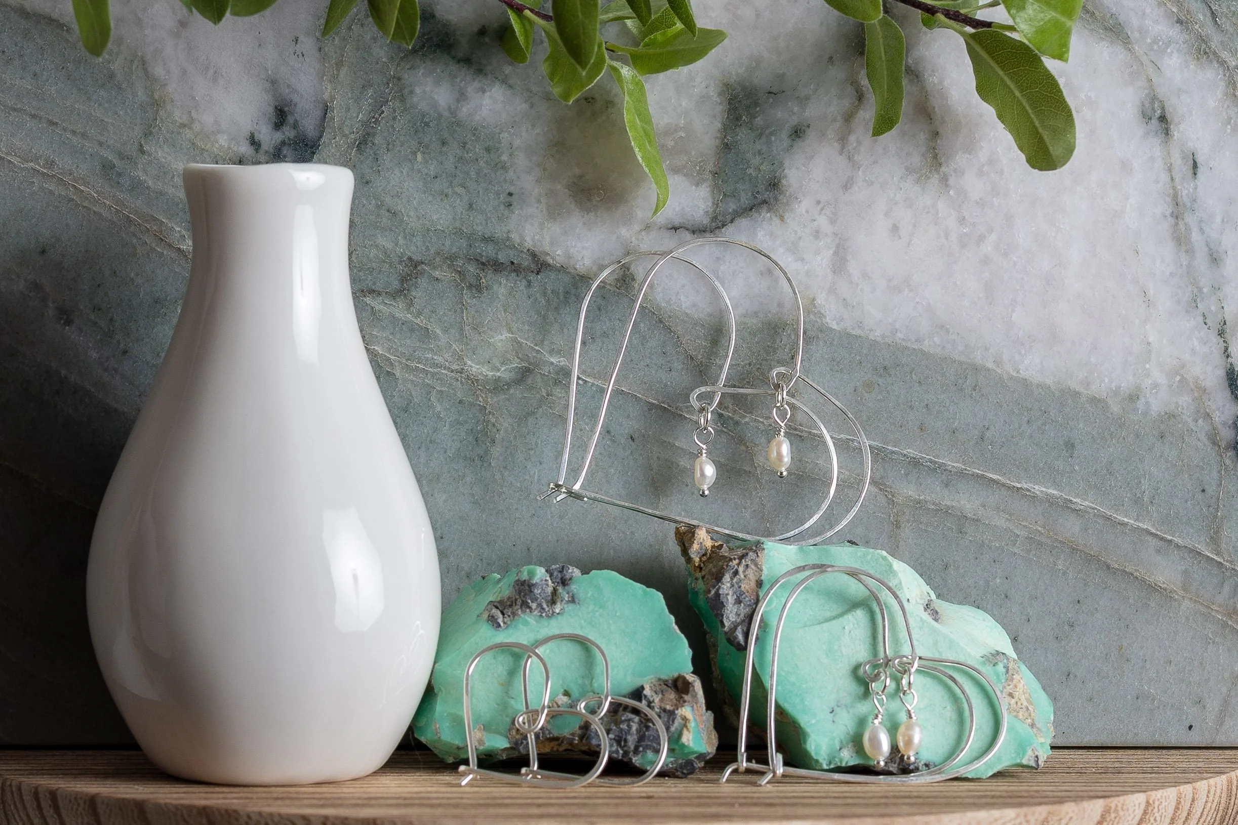 White ceramic vase and silver jewelry, including earrings with pearl drops, displayed on a wooden surface with turquoise and black stone accents, against a stone wall background with green leaves at the top.