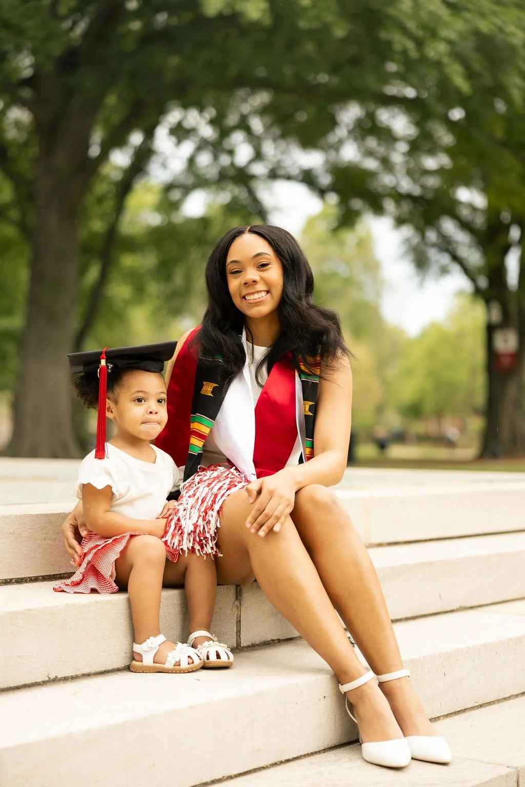 A Student-Mom from the University of Alabama graduates and sits on steps on campus with her baby by her side who is holding an Alabama game day shaker